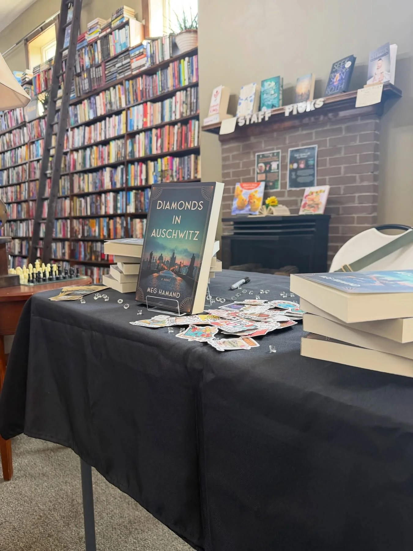 A table covered with a black cloth displays copies of the book 'Diamonds in Auschwitz' by Meg Hamand, along with scattered cards and small decorative items. Behind the table, a brick wall features a bookshelf with a variety of books and a sign labele