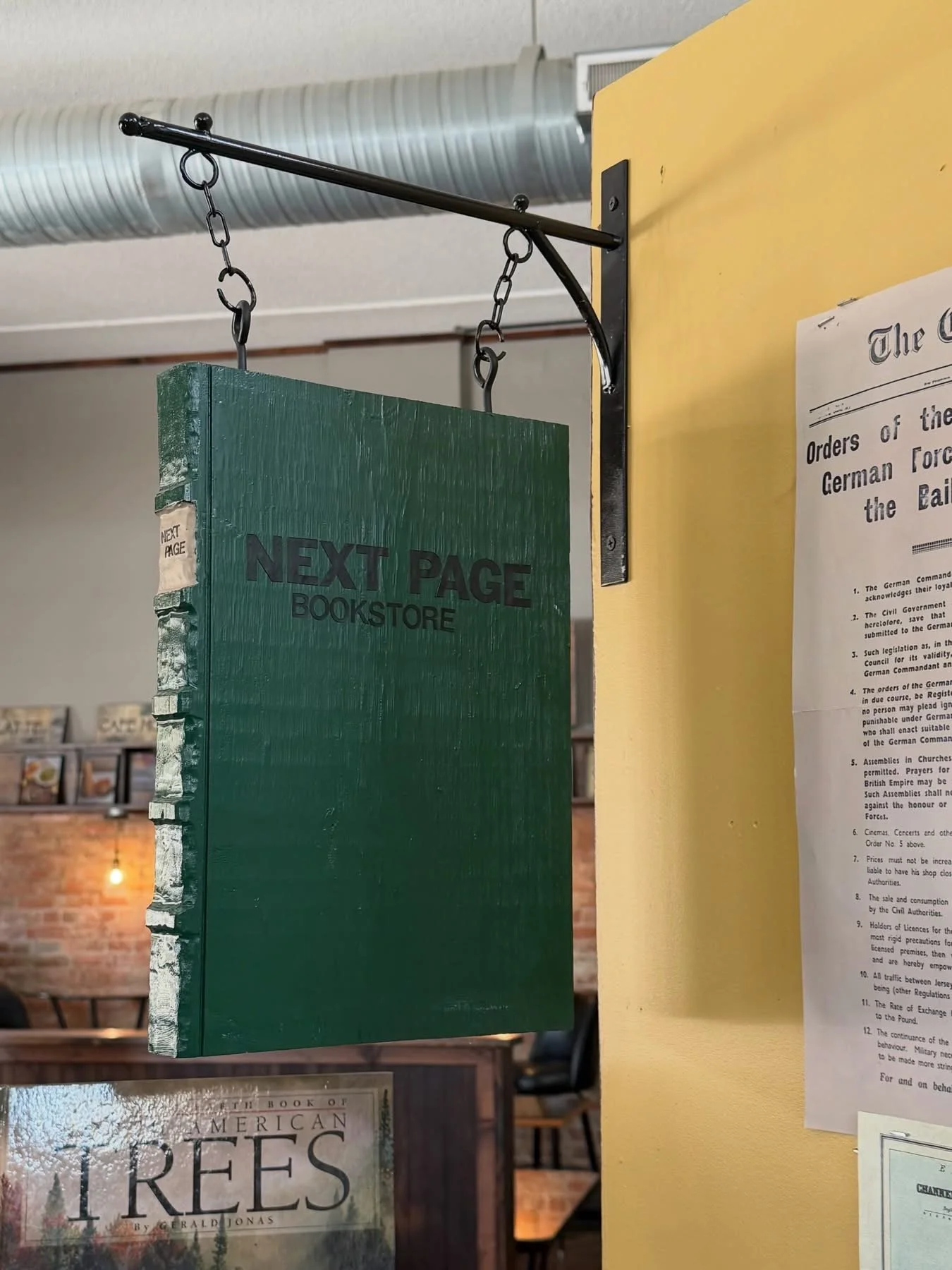 Green wooden sign for 'Next Page Bookstore' hanging from an iron hook in a bookstore with shelves of books in the background.