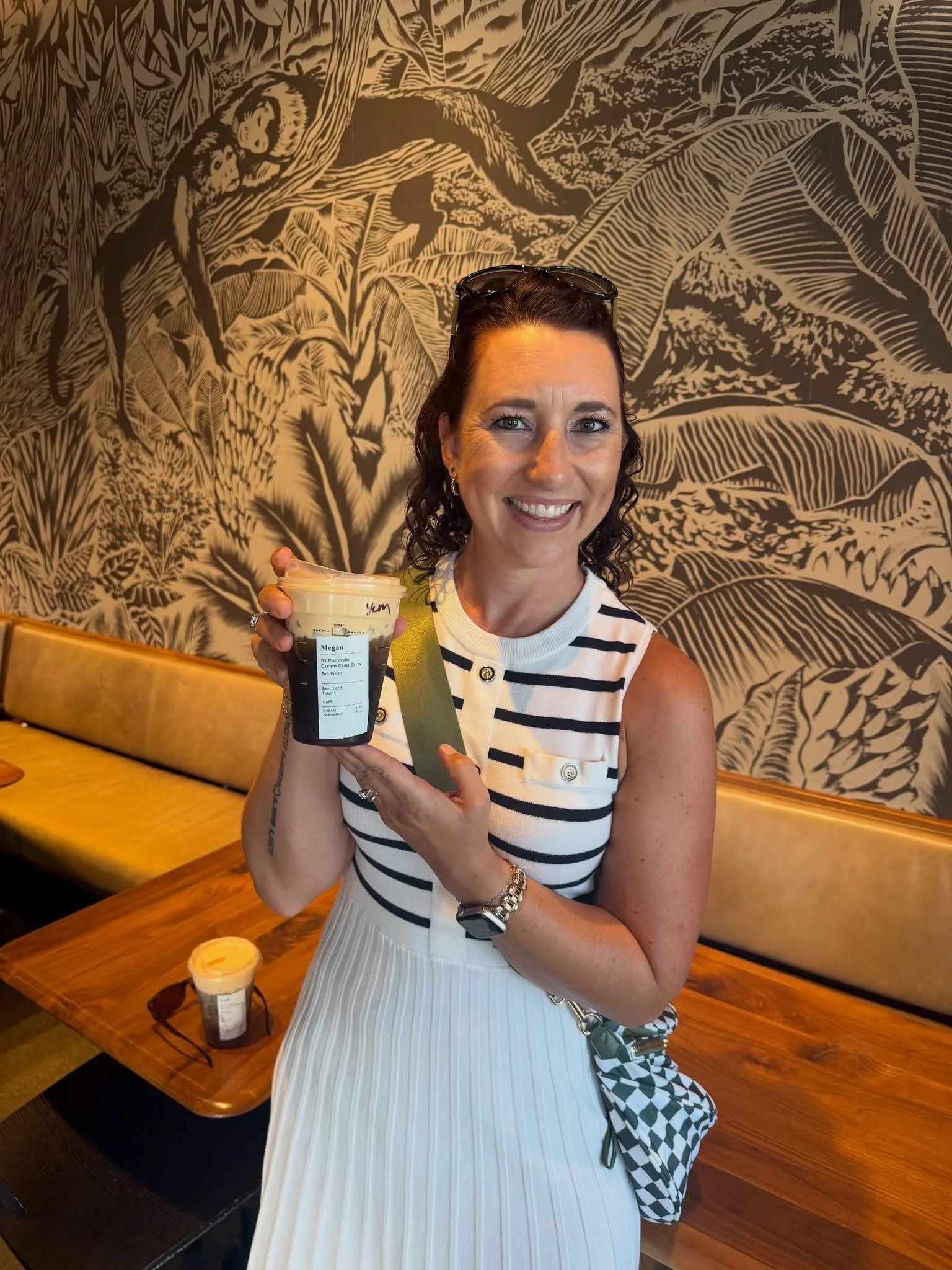 A woman with dark curly hair wearing a white sleeveless dress with black horizontal stripes, smiling, holding a plastic cup of coffee or tea, sitting in a café with wooden tables and a wall with jungle-themed artwork in the background.