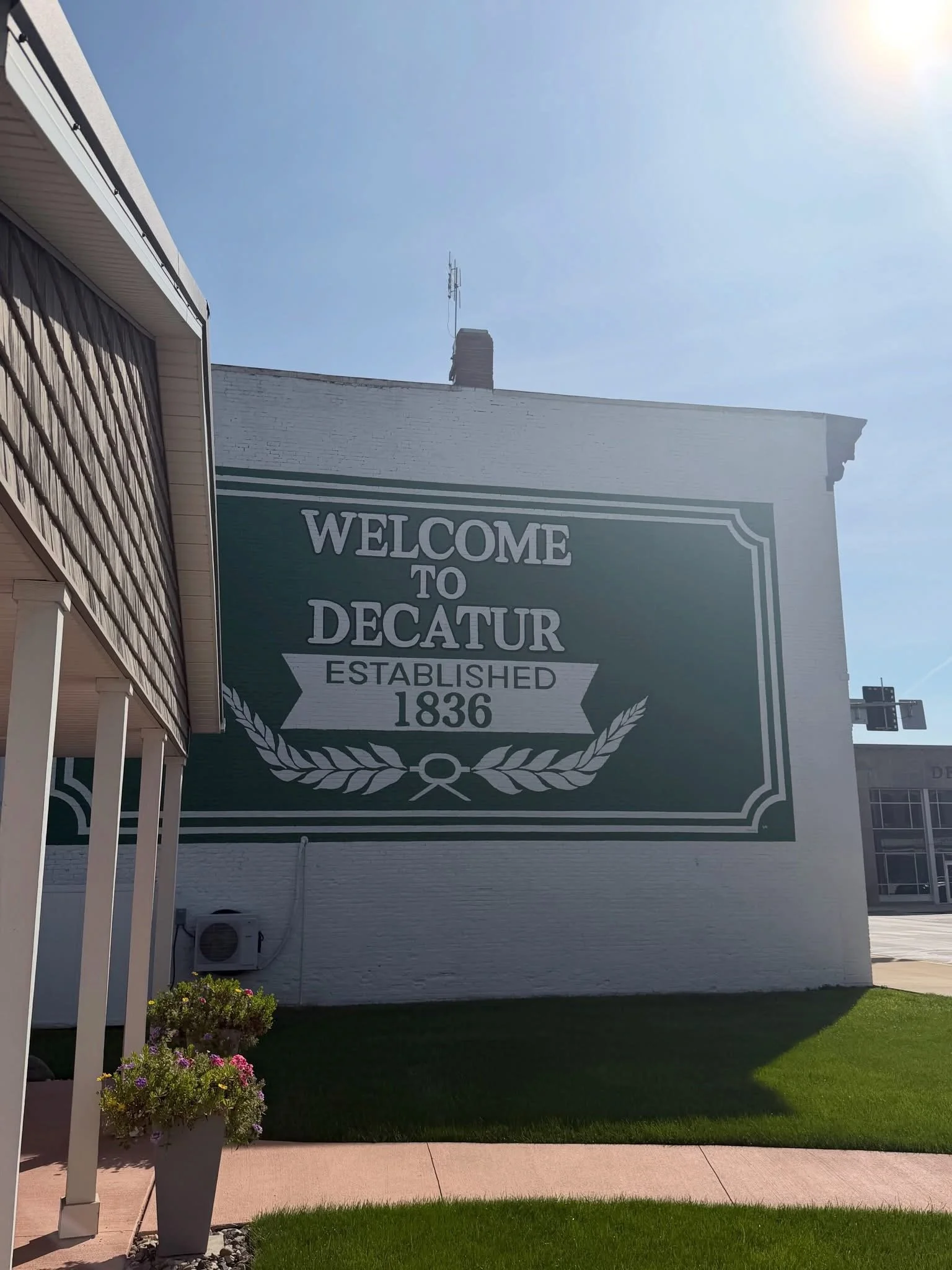 A large sign on a white brick building reads, "Welcome to Decatur. Established 1836." The building has a chimney with a communication antenna mounted on top. There is a well-maintained lawn with pink and white flowers in a gray pot, and a sidewalk in