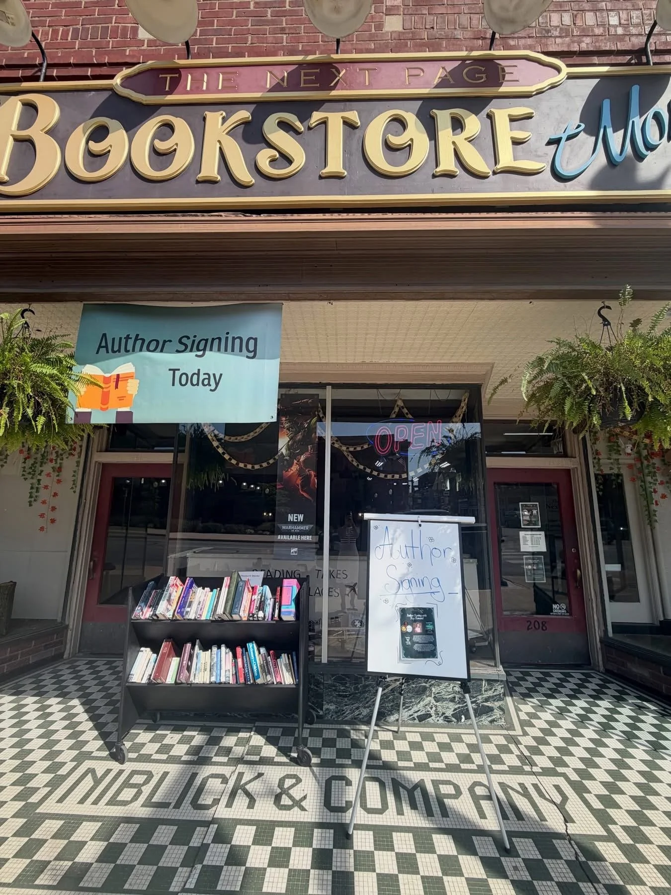 Exterior of a bookstore with a sign that says 'The Next Page Bookstore' and a banner advertising an author signing event today. There are books displayed outside on a shelf, a whiteboard with 'Author Signing' written on it, and a neon 'Open' sign in 