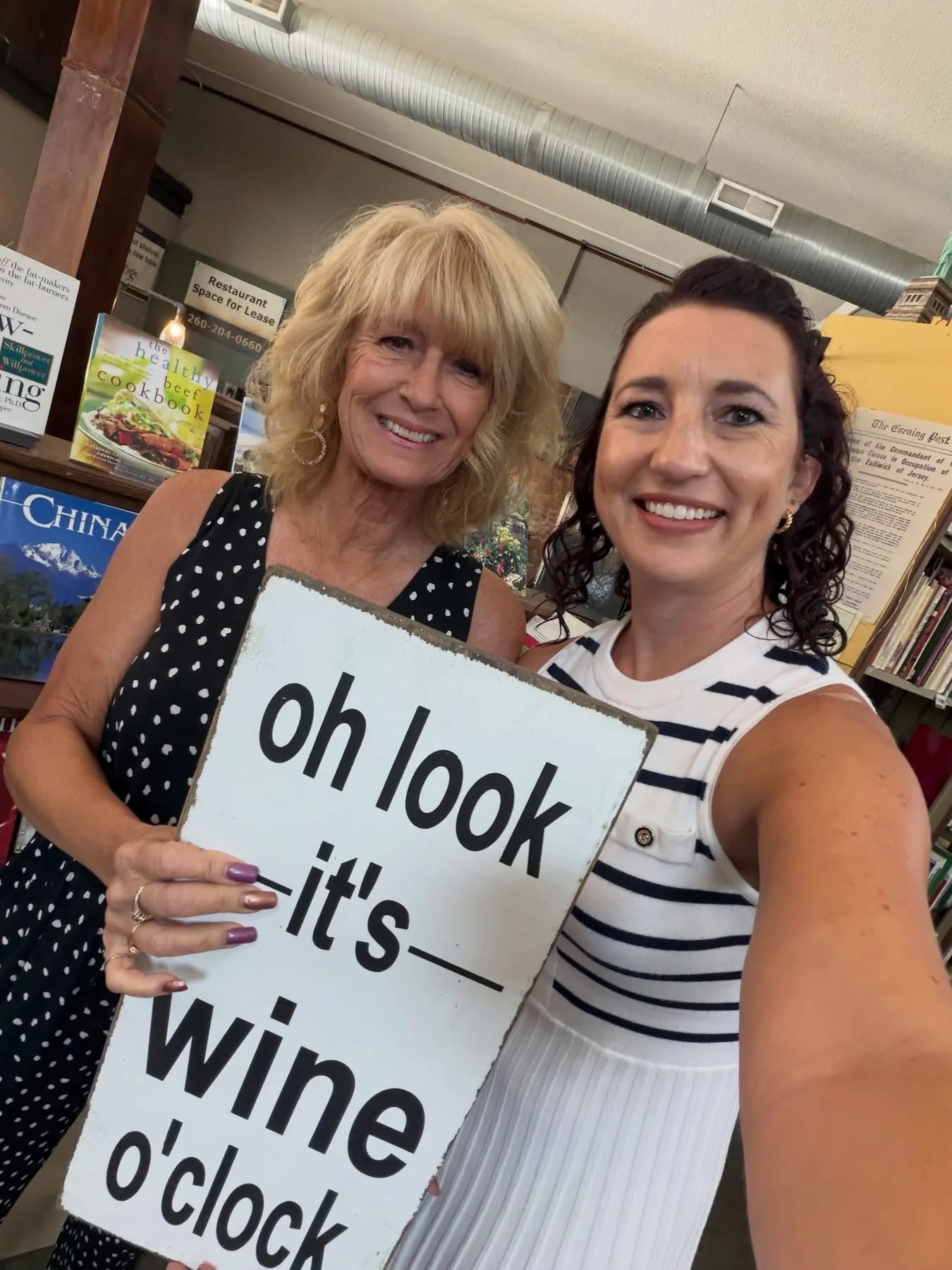 Two women smiling for a selfie in a bookstore, holding a sign that says "oh look it's" followed by partially obscured text. Books and posters can be seen in the background.