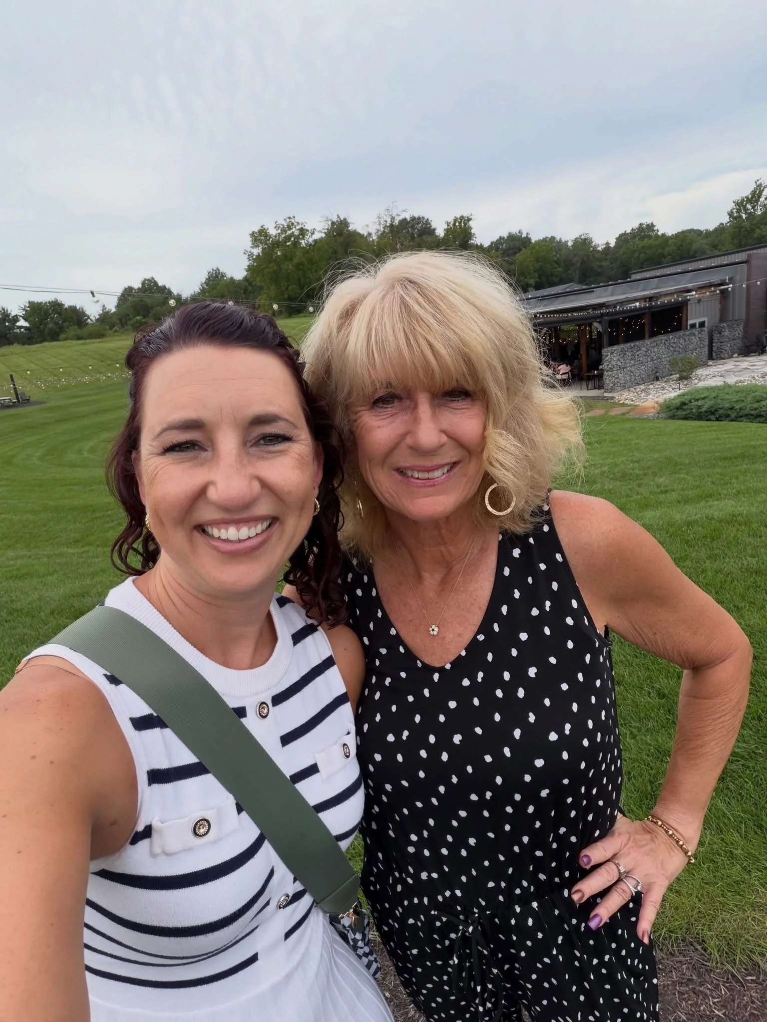 Two women smiling outdoors in front of a green lawn, trees, and a building with string lights, during daytime.