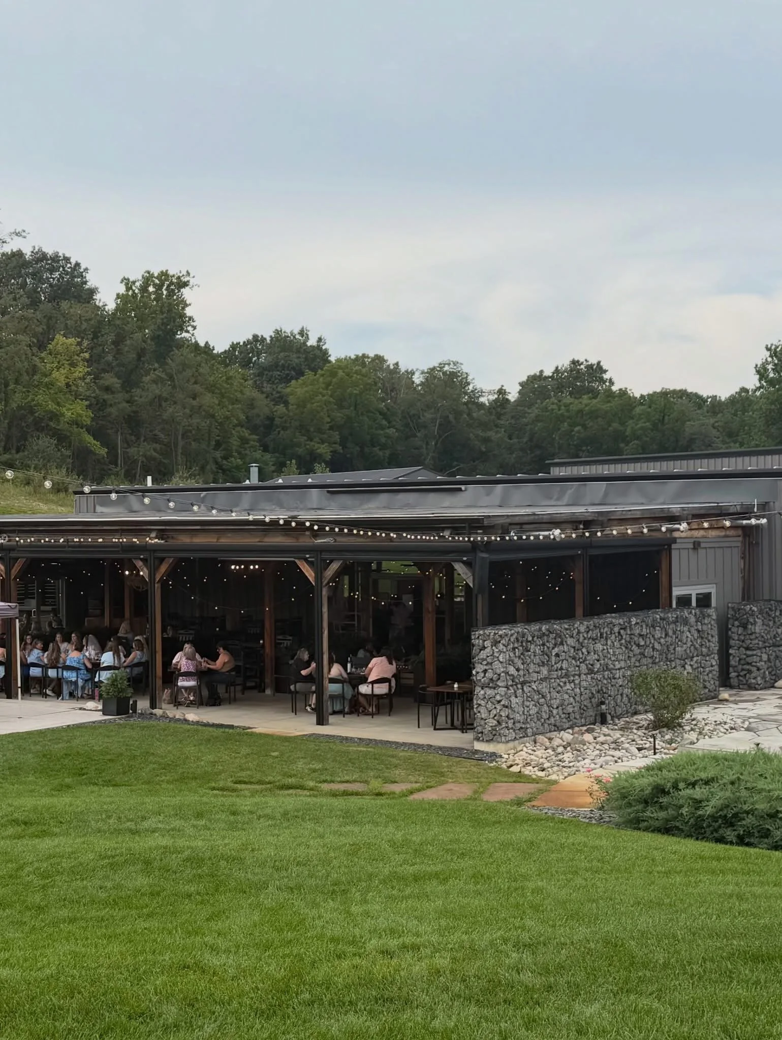 An outdoor patio with string lights, a grassy lawn, a building with string lights, trees, and a cloudy sky.
