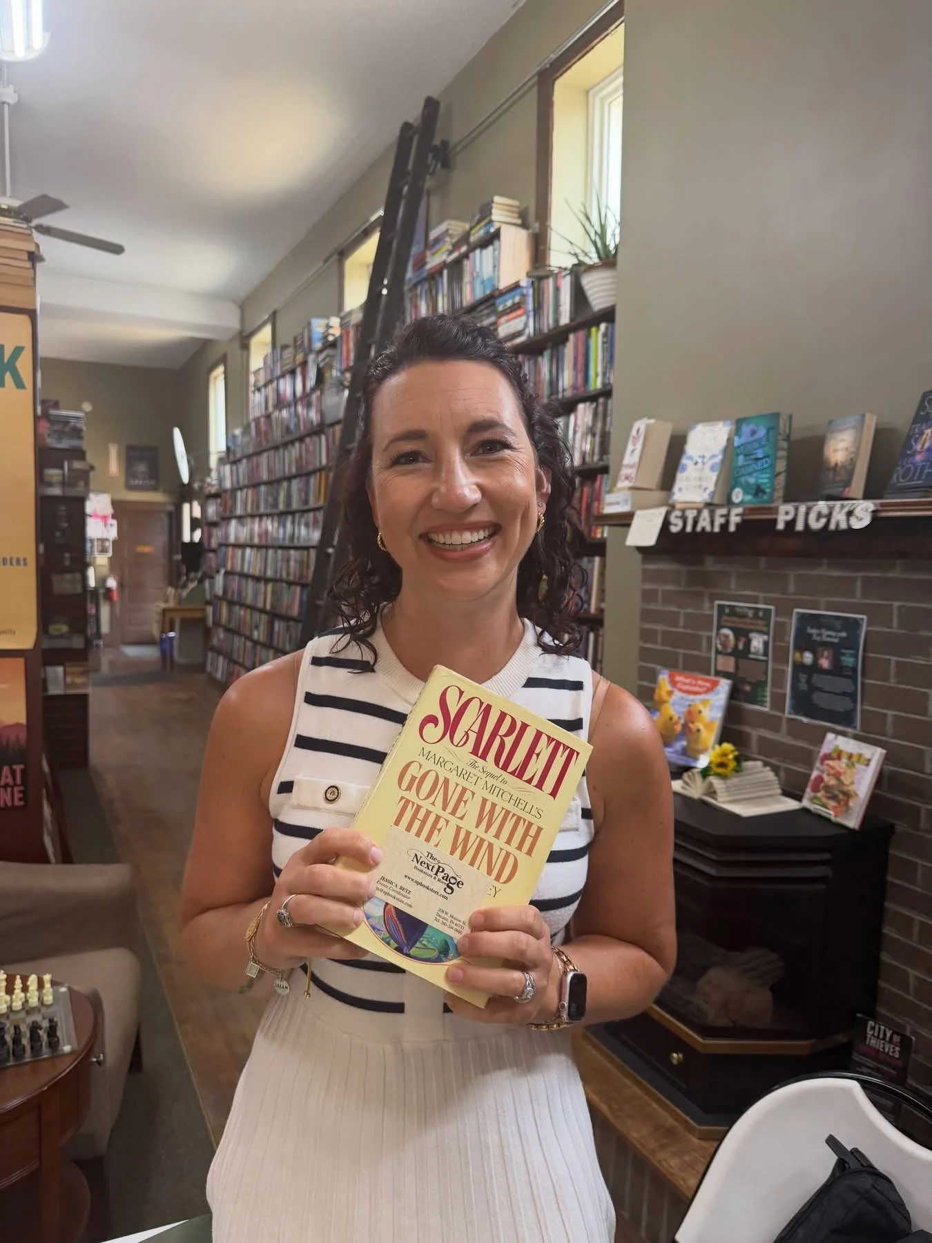 A smiling woman with dark curly hair holding a book titled 'Gone With the Wind' in a bookstore or library environment with bookshelves in the background.