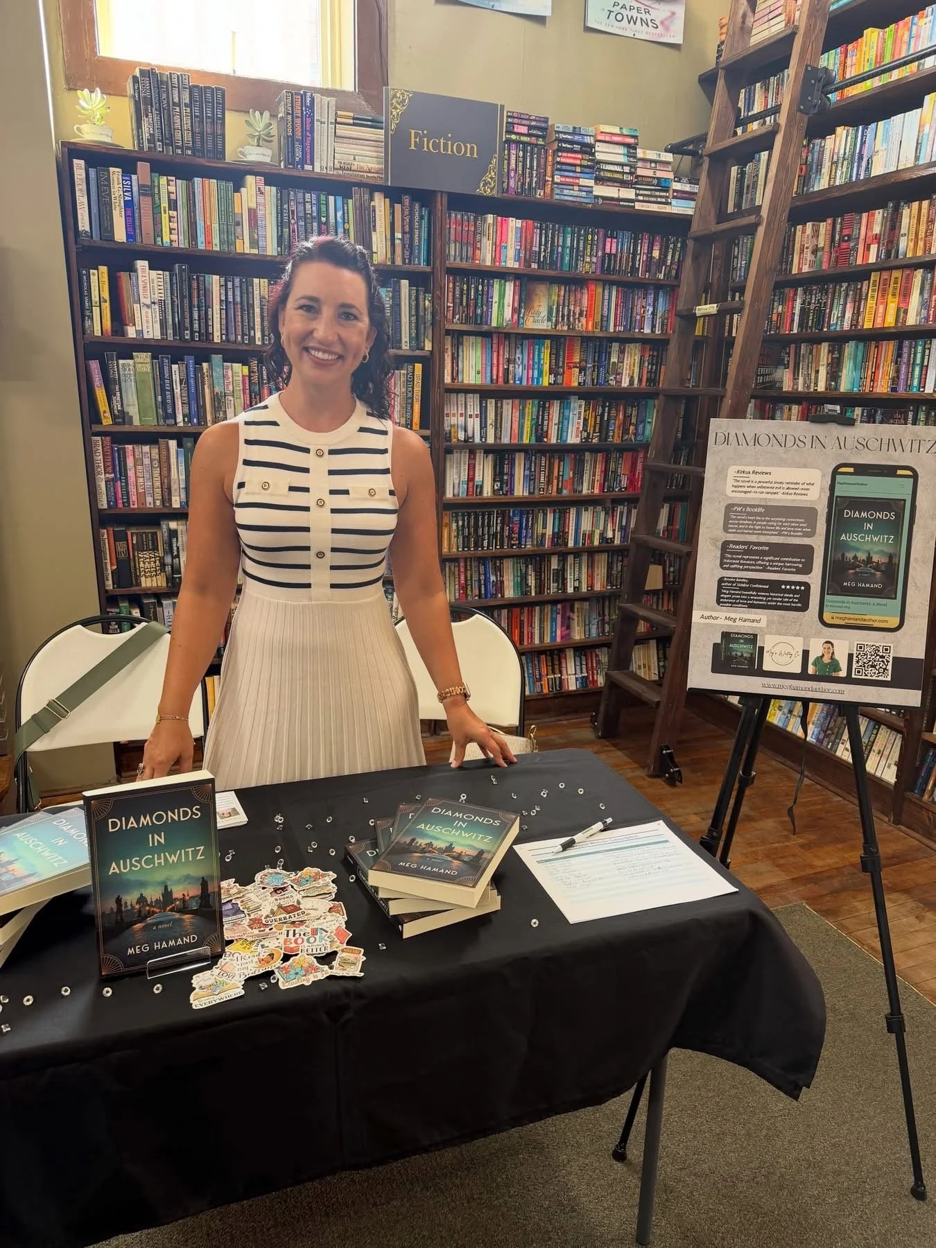 A woman smiling behind a table displaying her book titled "Diamonds in Auschwitz" at a book signing event in a bookstore. The table has copies of her book, promotional materials, and a sheet of paper. The background features tall bookshelves filled w