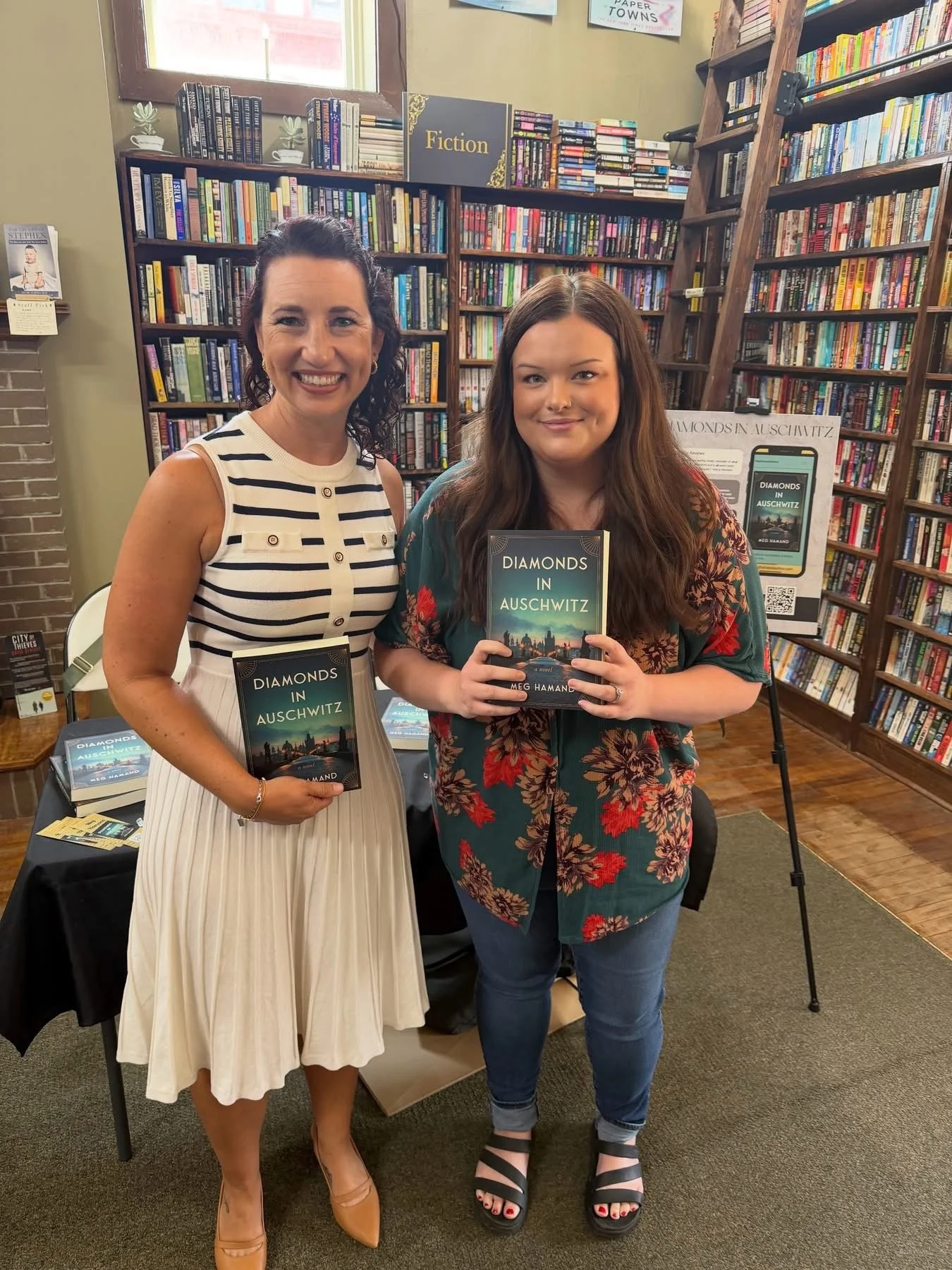Two women standing in a bookstore, holding copies of a book titled "Diamonds in Auschwitz." The woman on the left is wearing a sleeveless white dress with black stripes, beige high heels, and a bracelet on her right wrist. The woman on the right is w