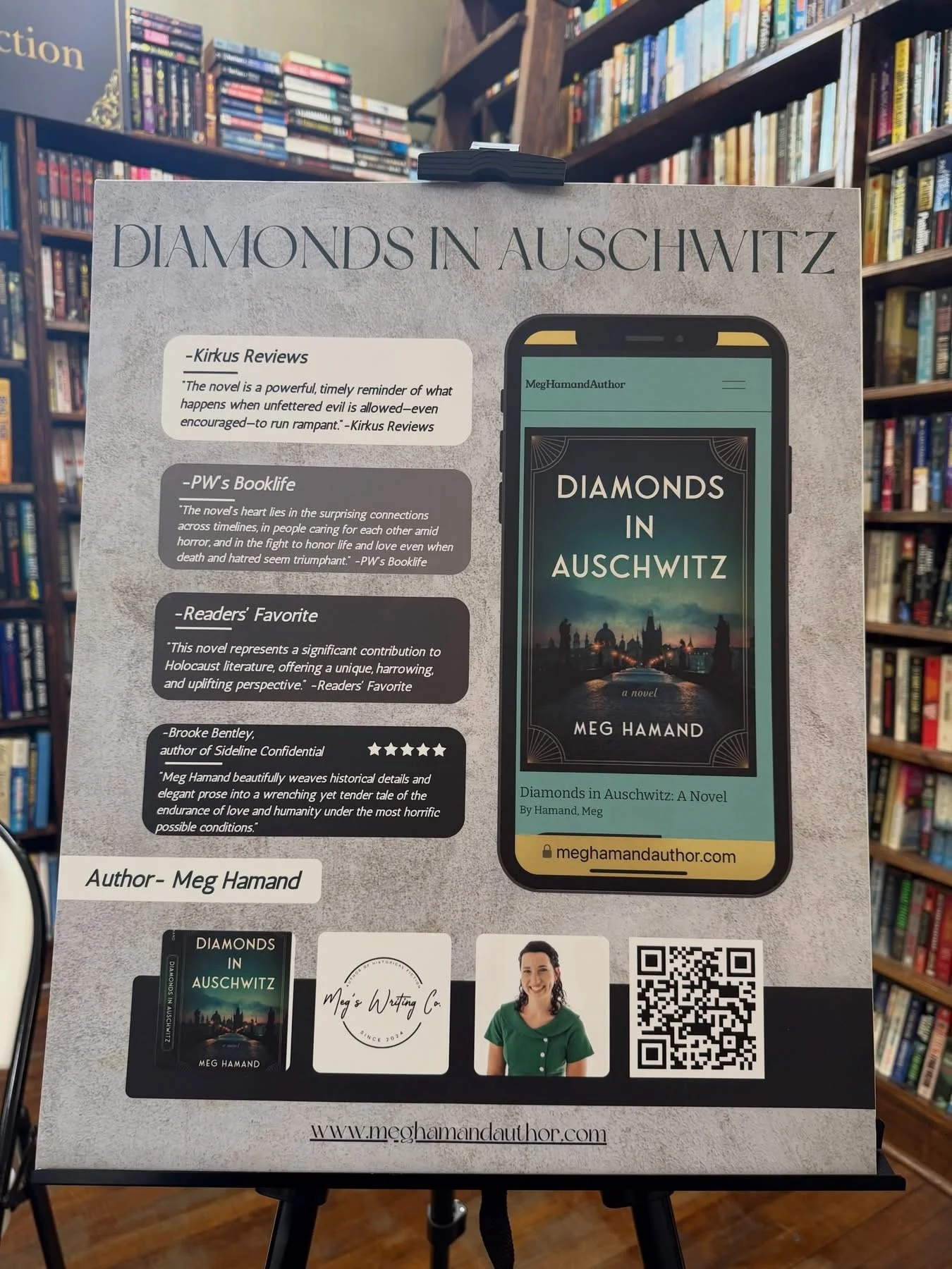Bookstore display promoting the novel "Diamonds in Auschwitz" by Meg Hamand. The display includes reviews, author information, a QR code, and an image of the book cover on a phone screen, set against bookshelves in the background.