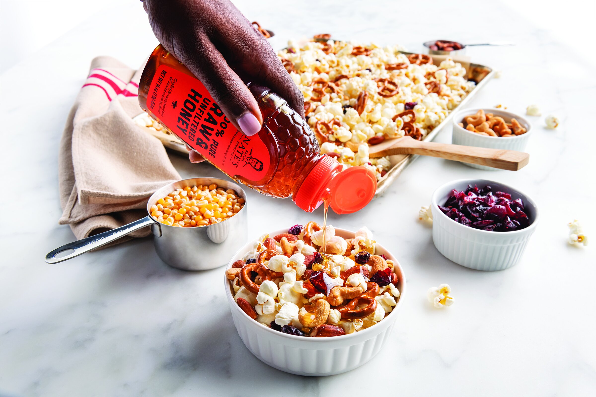 Dramatic photo of Nature Nate's Honey being drizzled over a snack bowl of popcorn, nuts, and pretzels.