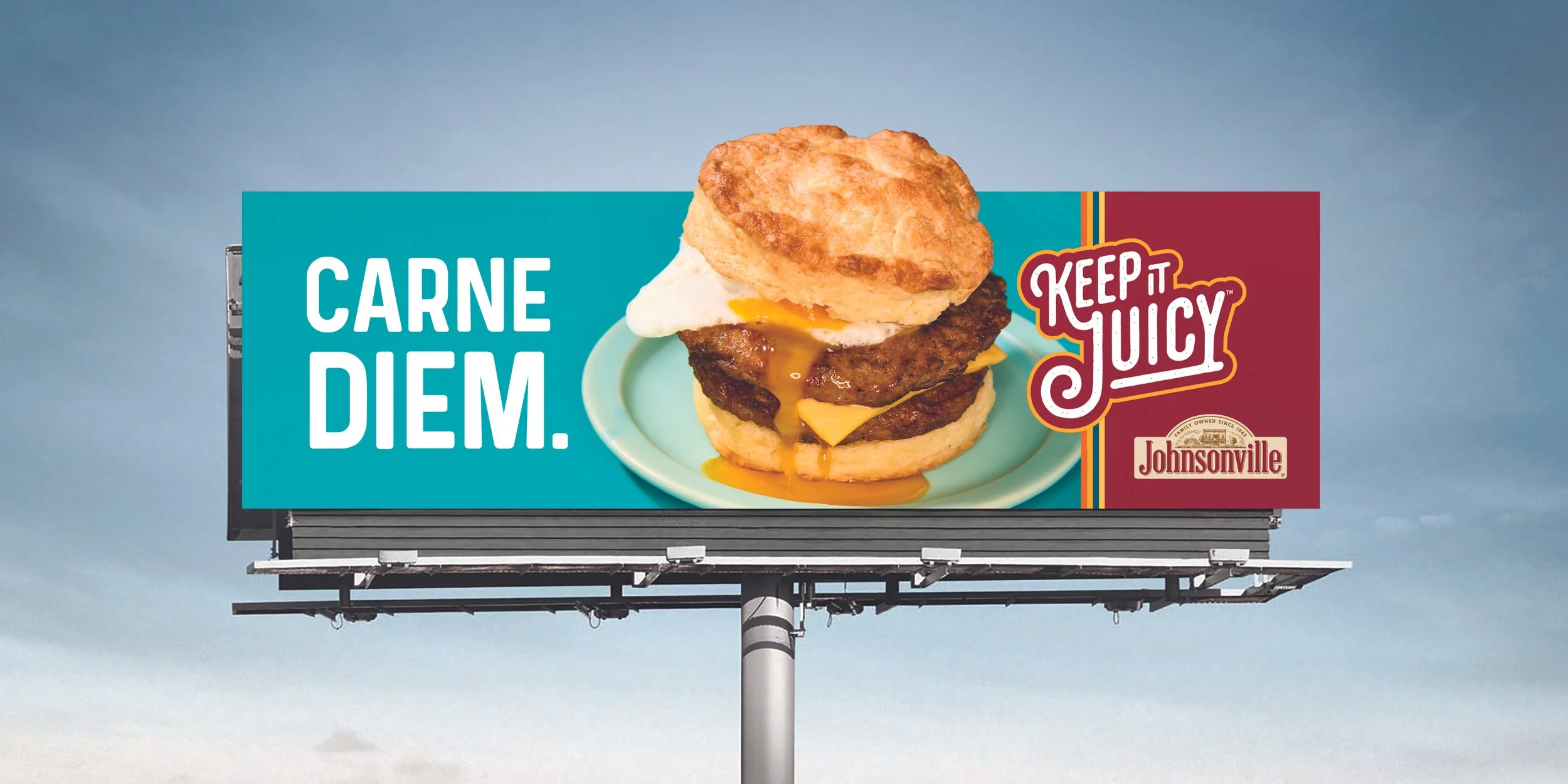 An outdoor board for Johnsonville Sausage featuring a photo of a sausage biscuit and a headline that reads, "Carne Diem." The "Keep It Juicy" logo appears on the right side of the ad.