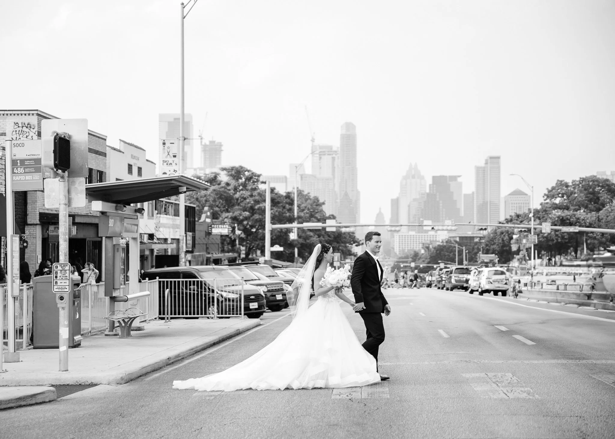 A bride and groom walking hand in hand across a city street, with the bride in a white wedding dress and veil, holding a bouquet, and the groom in a dark tuxedo, in an urban setting with tall buildings and parked cars.