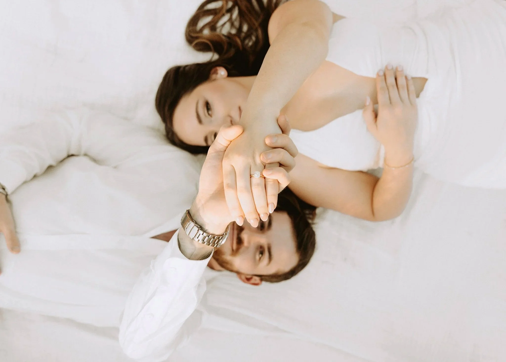 A woman with long brown hair lying on a white bed, holding her arm while a person out of frame touches her arm. The woman wears a black top, a ring, and a watch.