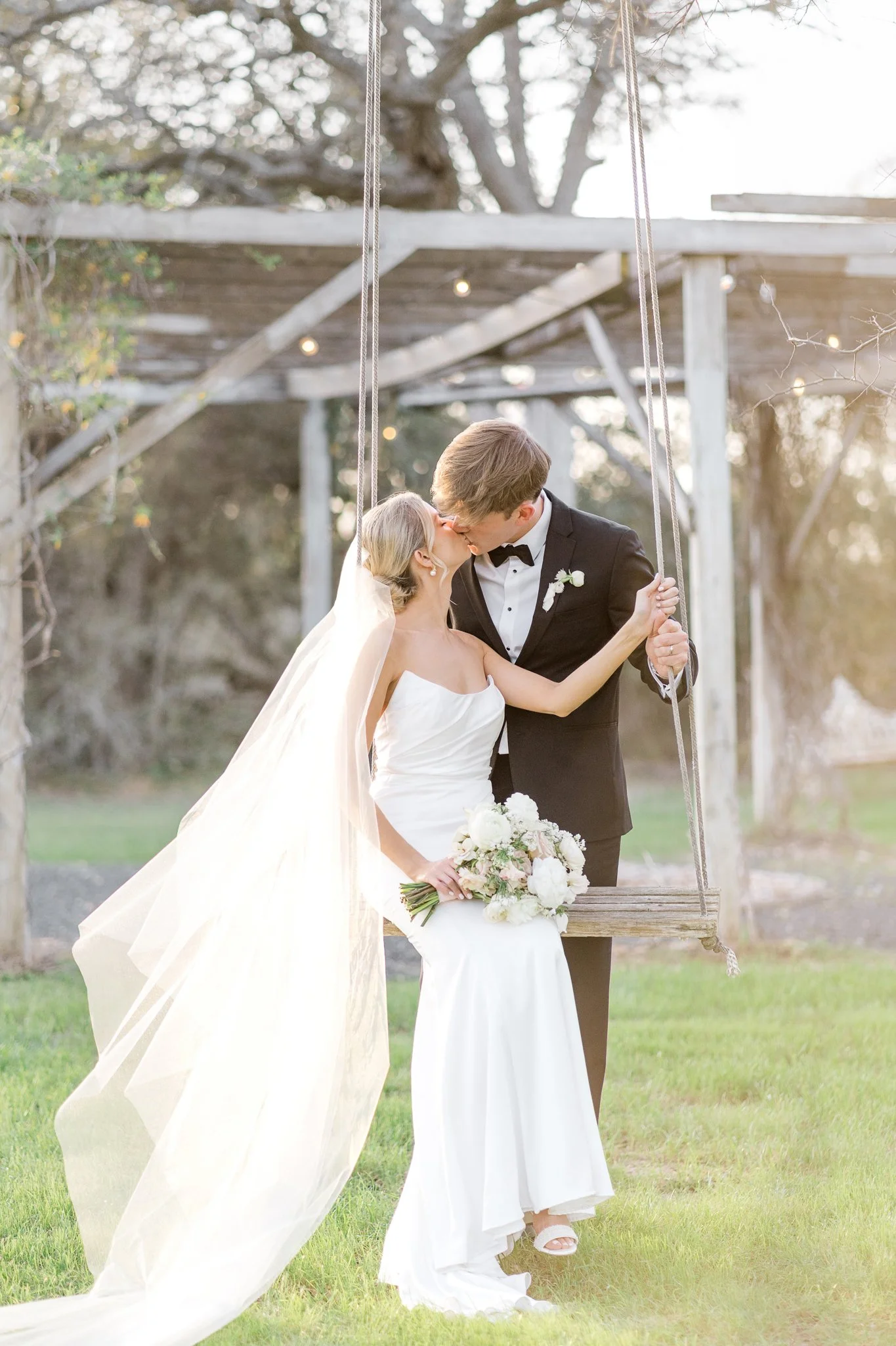 A bride and groom share a kiss outdoors, with the bride holding a bouquet of flowers, the bride wearing a white dress and the groom in a black tuxedo, sitting on a swing.