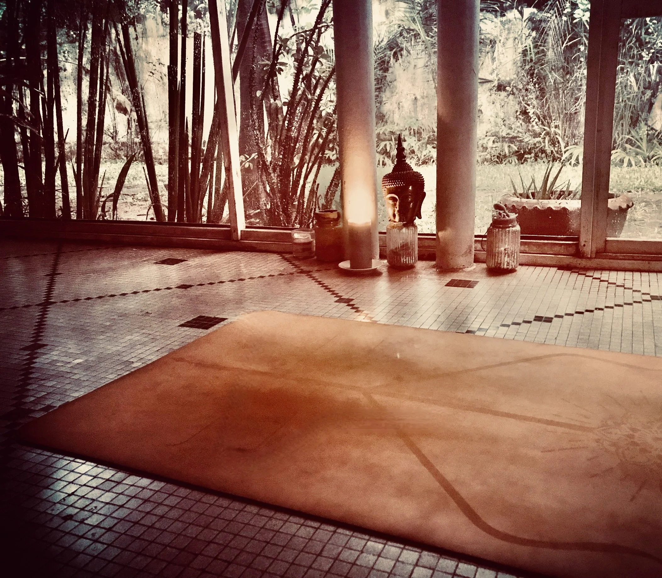 Indoor space with a yoga mat on a tiled floor near large windows overlooking a garden with lush greenery. Decorative items, including a Buddha statue and potted plants, are placed by the window.