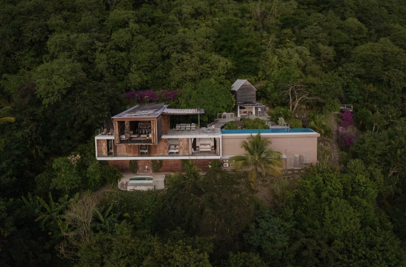Interior of a tropical-style hotel room with a thatched roof, wooden accents, a canopy bed with white linens, a separate bedroom with a window seat, and a bathroom with a round mirror in the background.