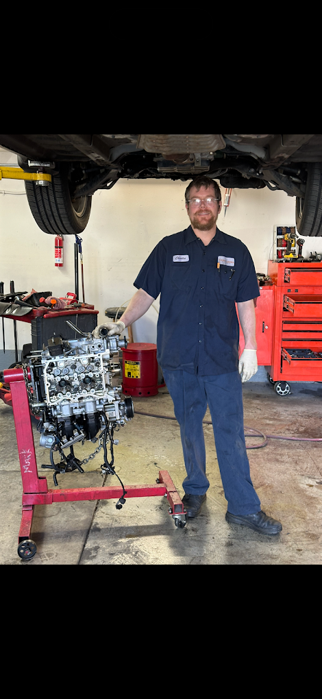 Automotive mechanic standing in an auto repair shop beneath a lifted vehicle, holding an engine on a red engine stand.