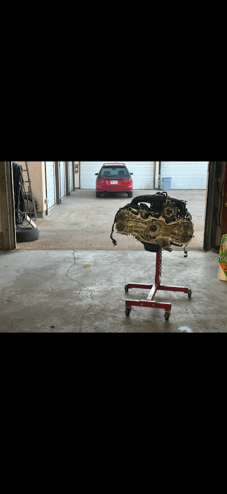 Car engine mounted on a red engine stand inside a garage, with a driveway and a red car visible outside.