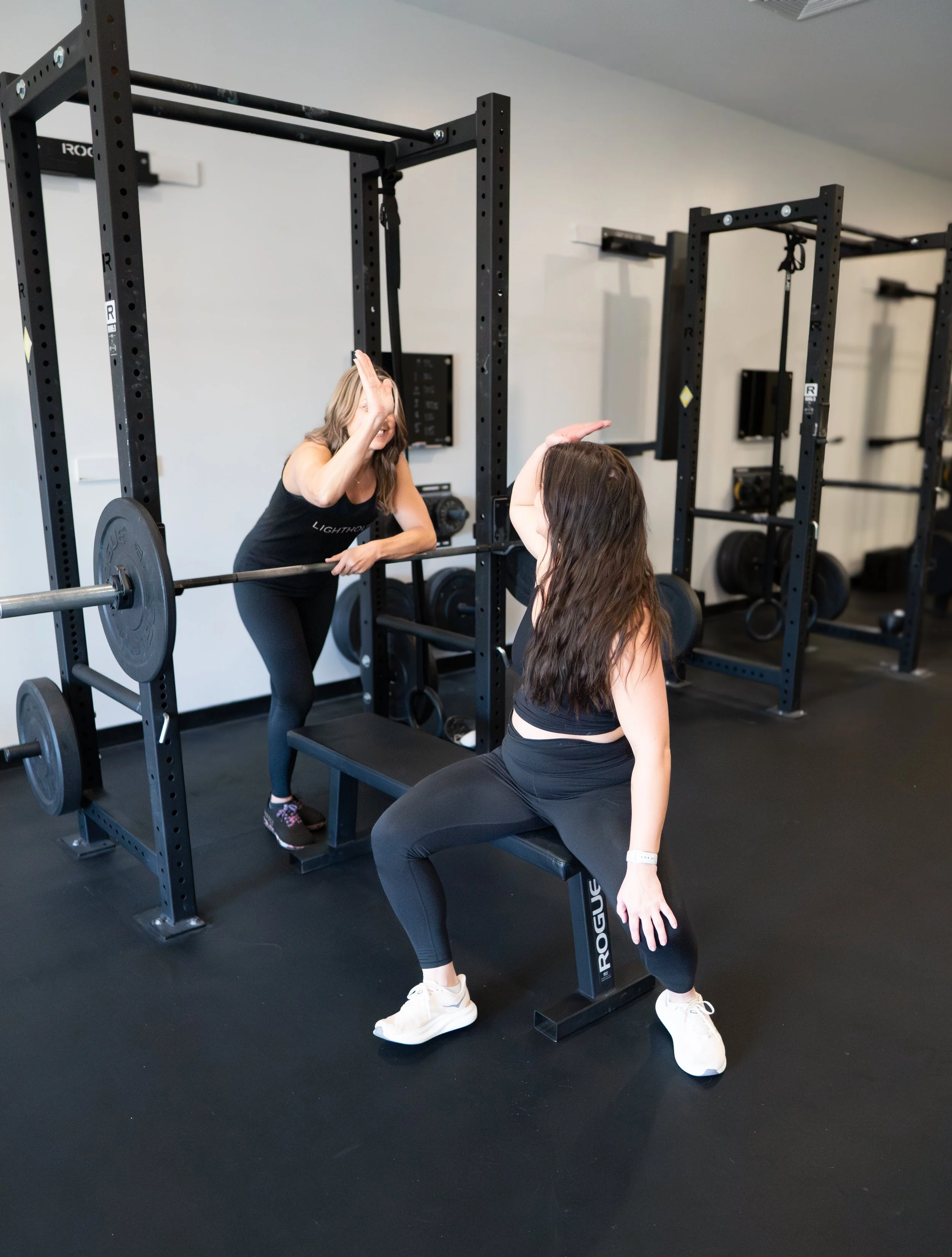 Two women in black athletic wear practicing high fives in a gym, with weight racks, weights, and a bench in the background.