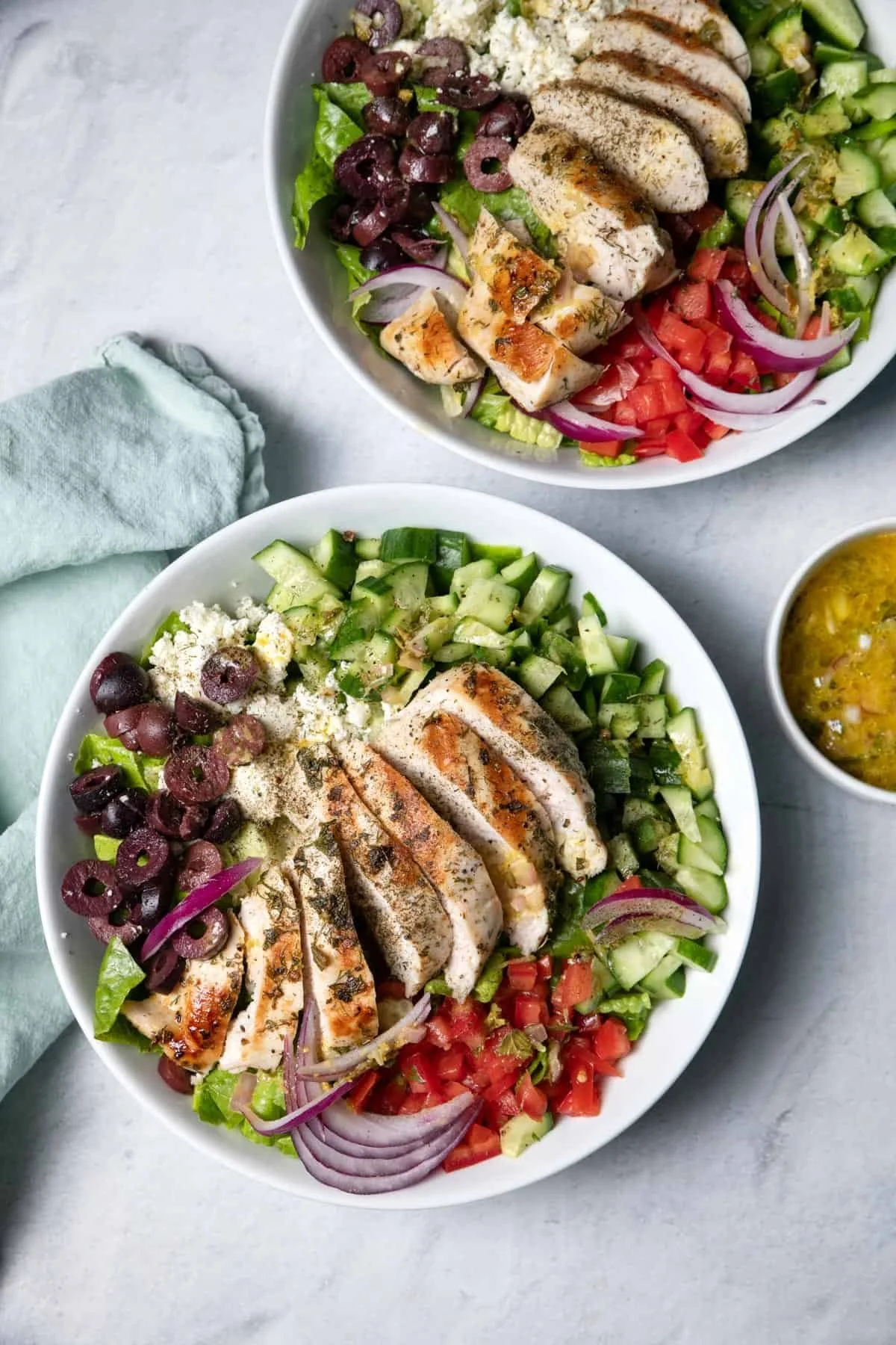 Two bowls of grilled chicken salad with tomatoes, cucumbers, red onions, shredded cheese, and black olives on a white surface, with a small bowl of salad dressing nearby.