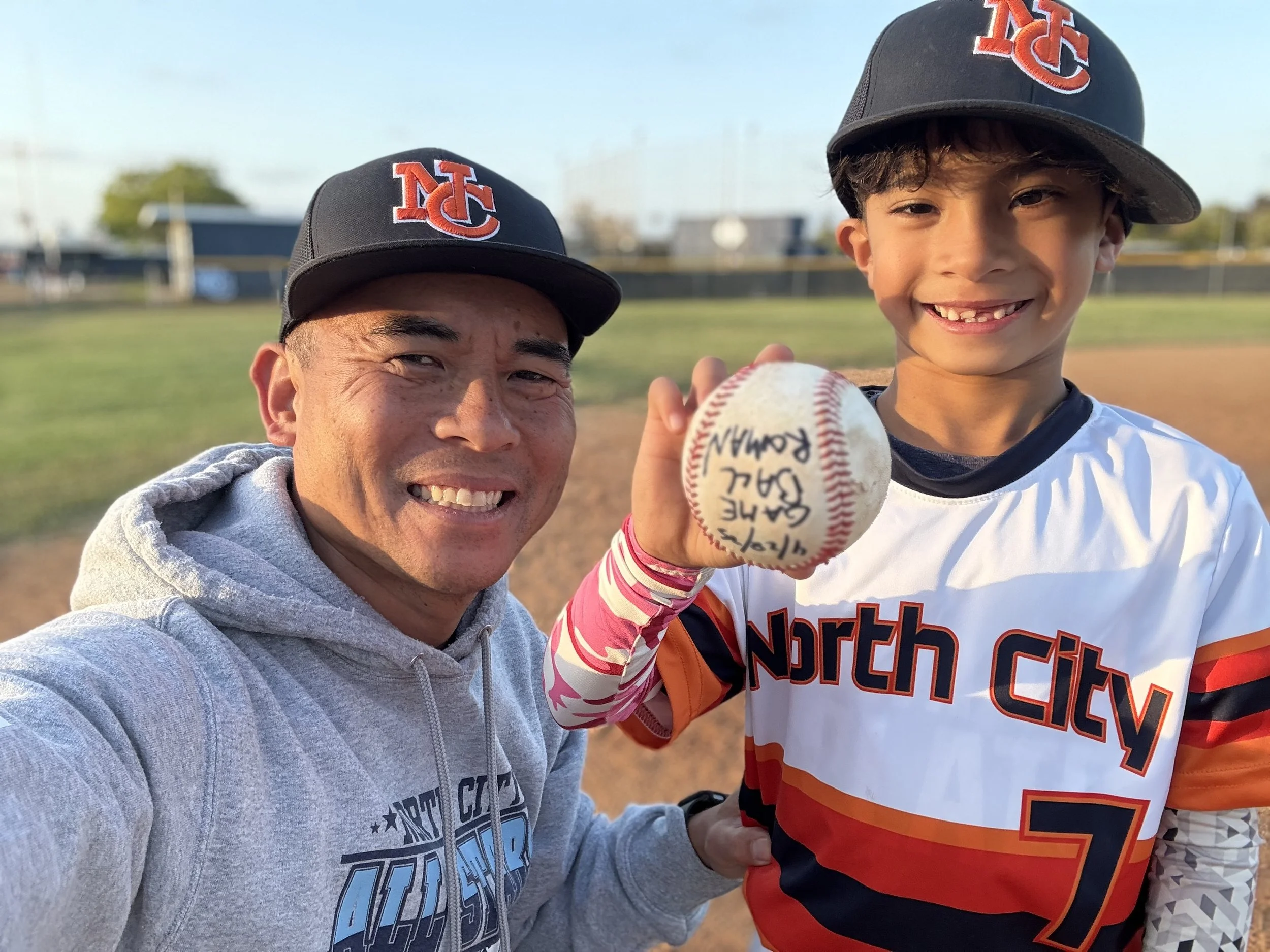 Boy is holding a gameball he earned during the game with a big smile next to his his coach/father.
