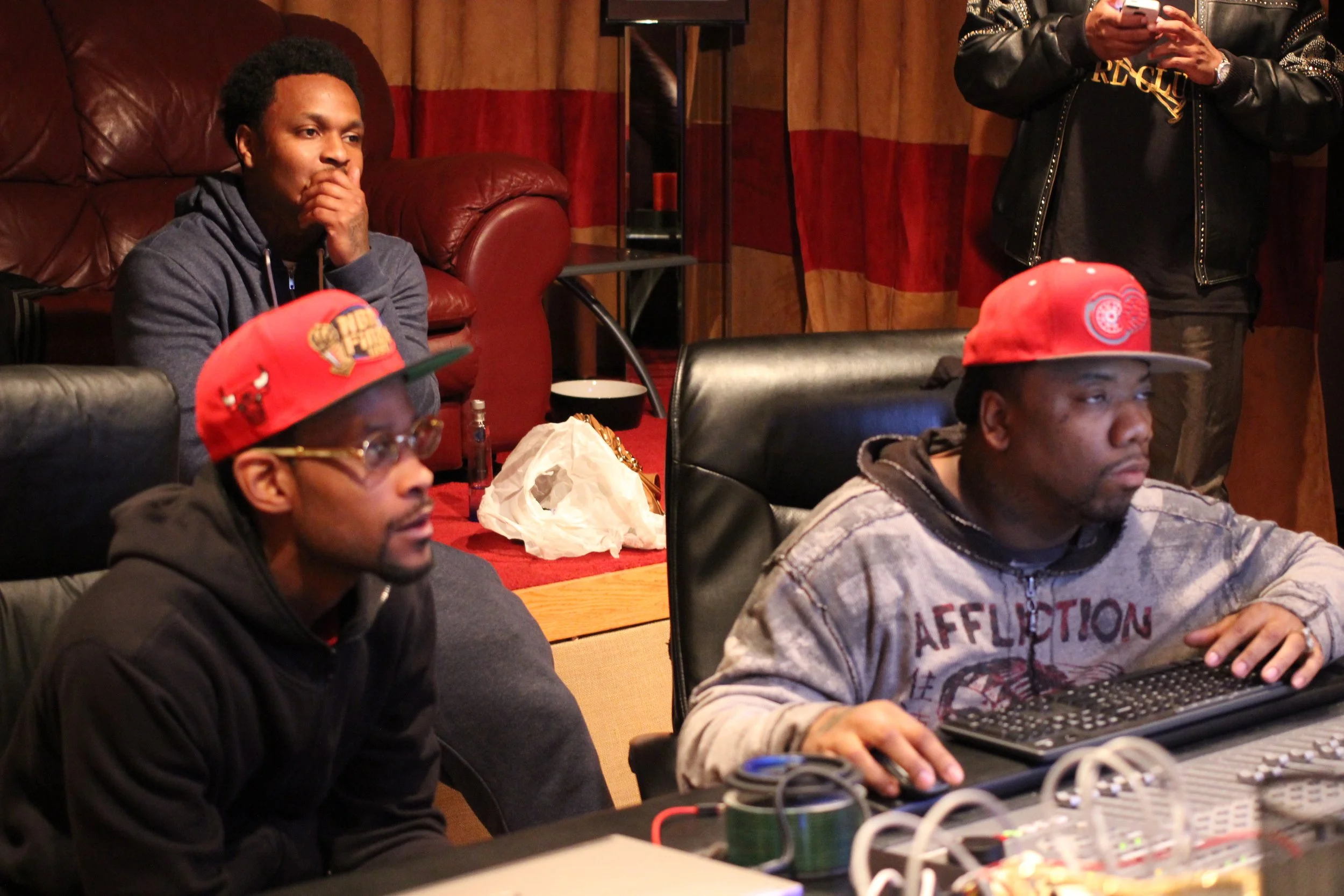 Three men in a studio or office setting, two wearing red Detroit Pistons caps, focusing on a computer screen, with equipment and a person in the background using a phone.