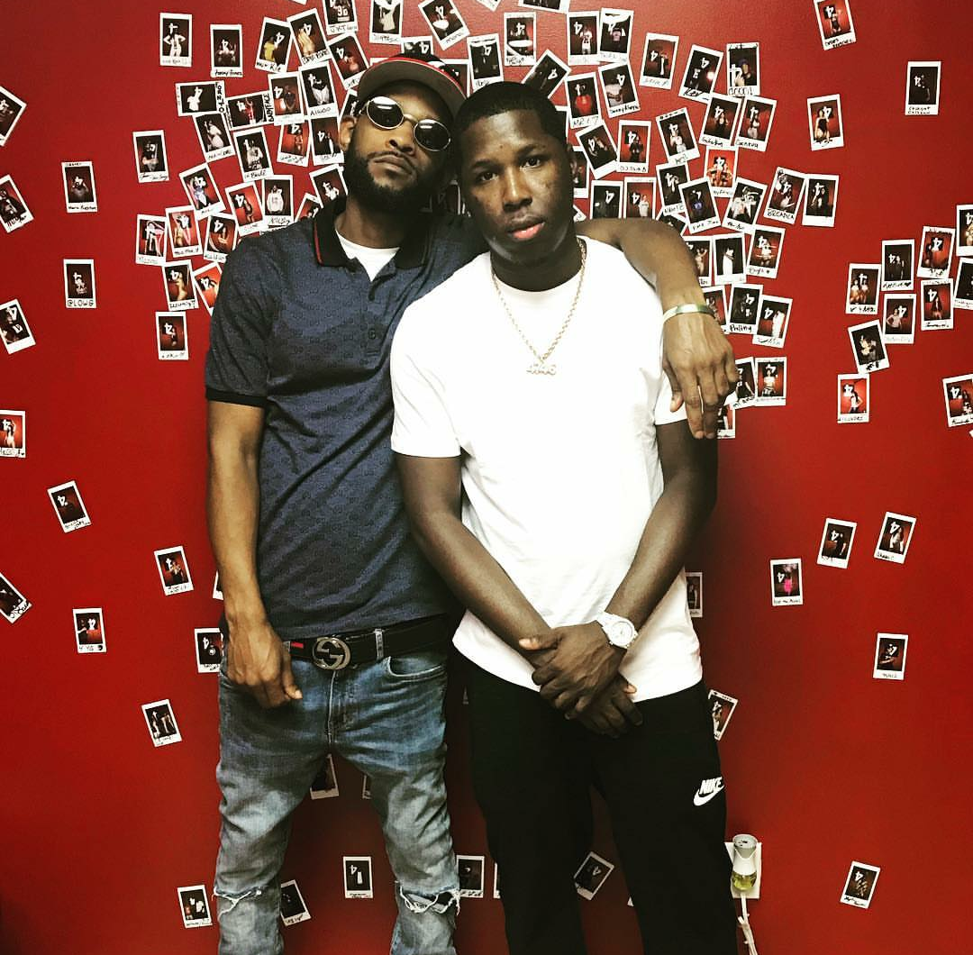 Two young men standing close together in front of a red wall decorated with numerous small photographs. The man on the left wears sunglasses, a black polo shirt, and ripped jeans, while the man on the right wears a white t-shirt, black Nike pants, and a gold chain, with his arm around the other man's shoulder.