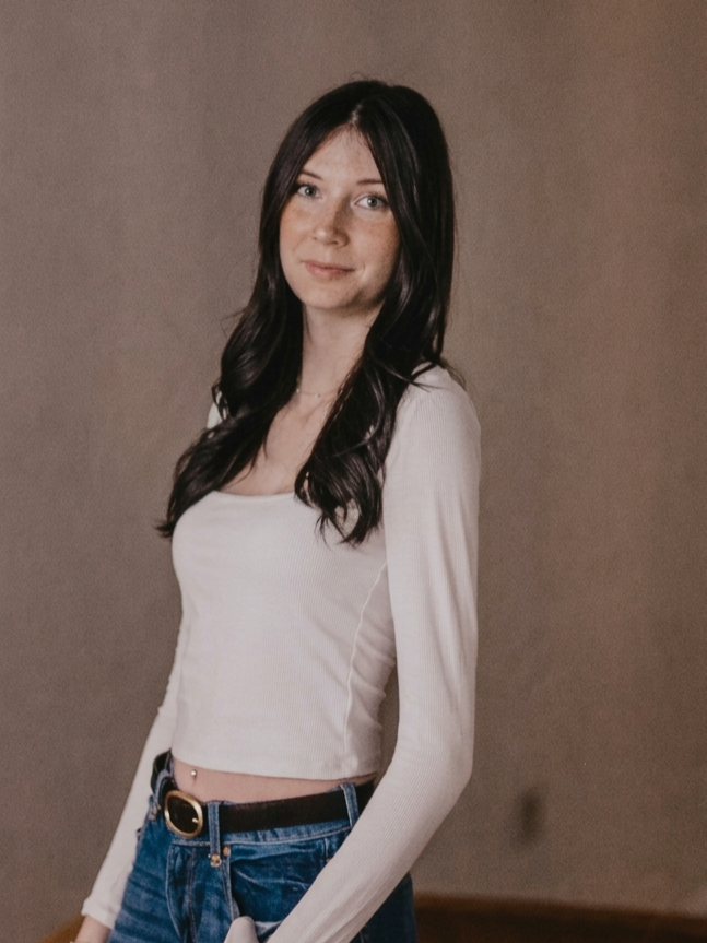 Young woman with long dark hair wearing a white long-sleeve crop top and blue jeans standing against a plain beige background.