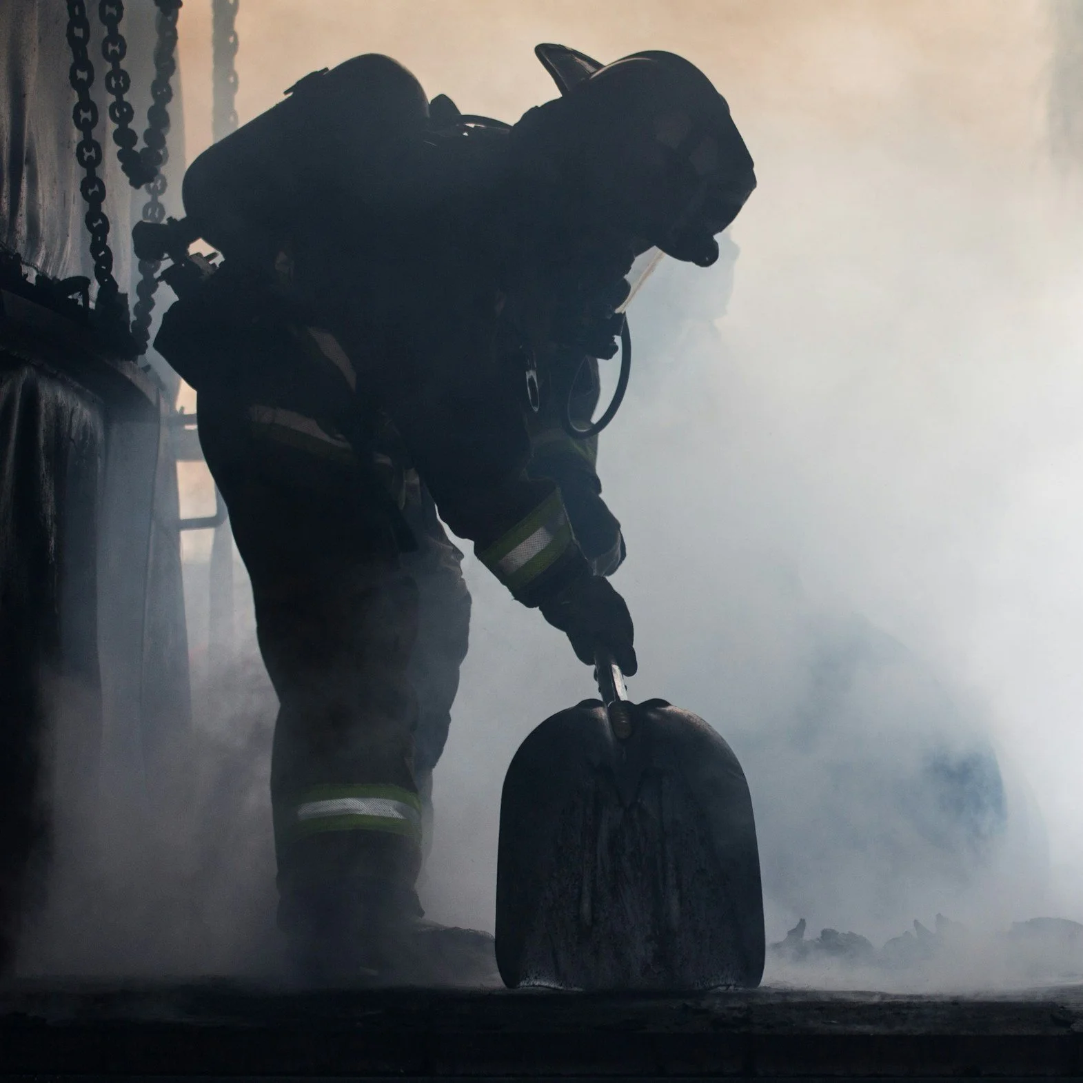 Firefighter in full gear, holding a tool, amid smoke in a smoky environment.