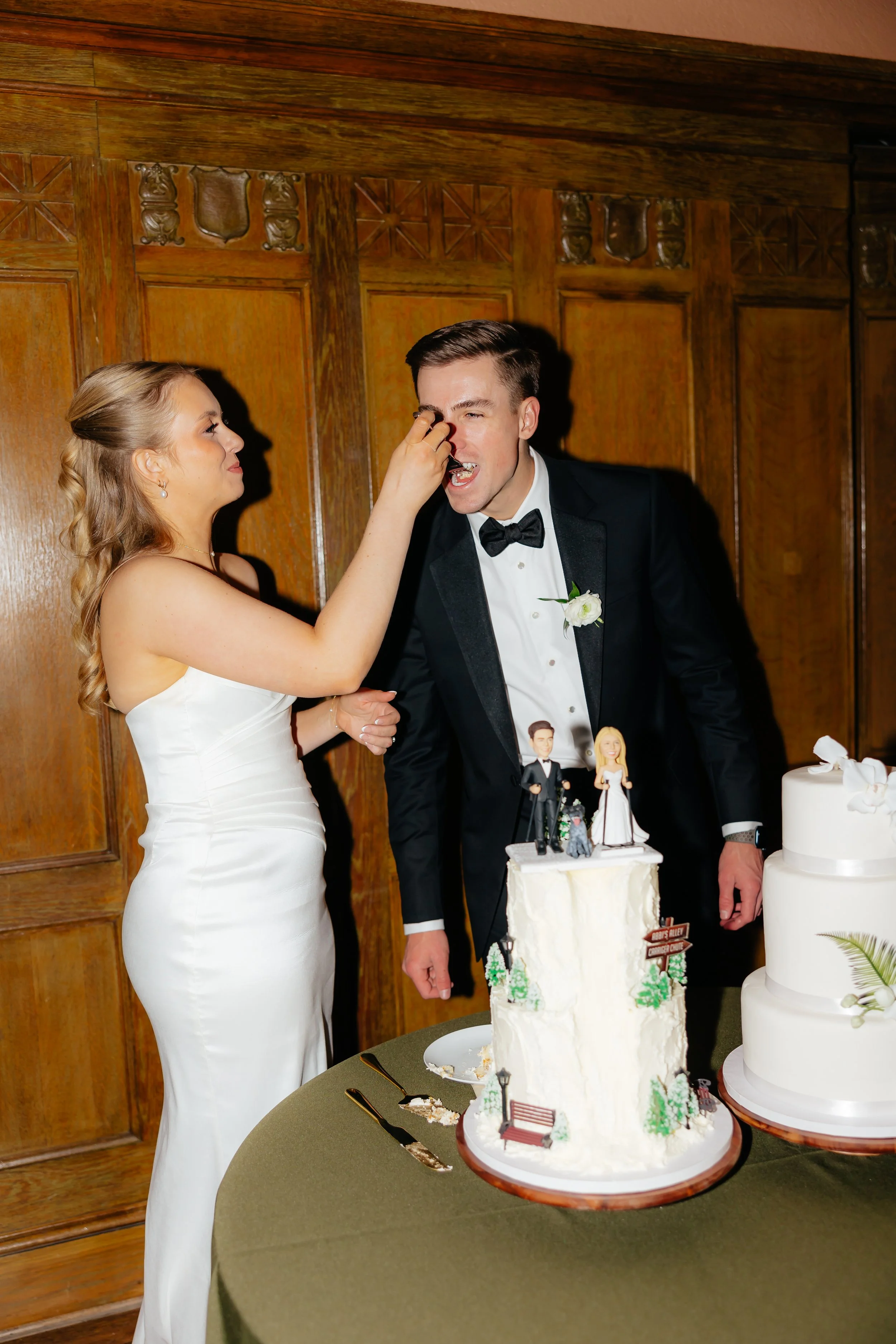 Bride and groom at wedding cake cutting celebration, with the bride feeding the groom a piece of cake, standing in front of wooden wall panels.