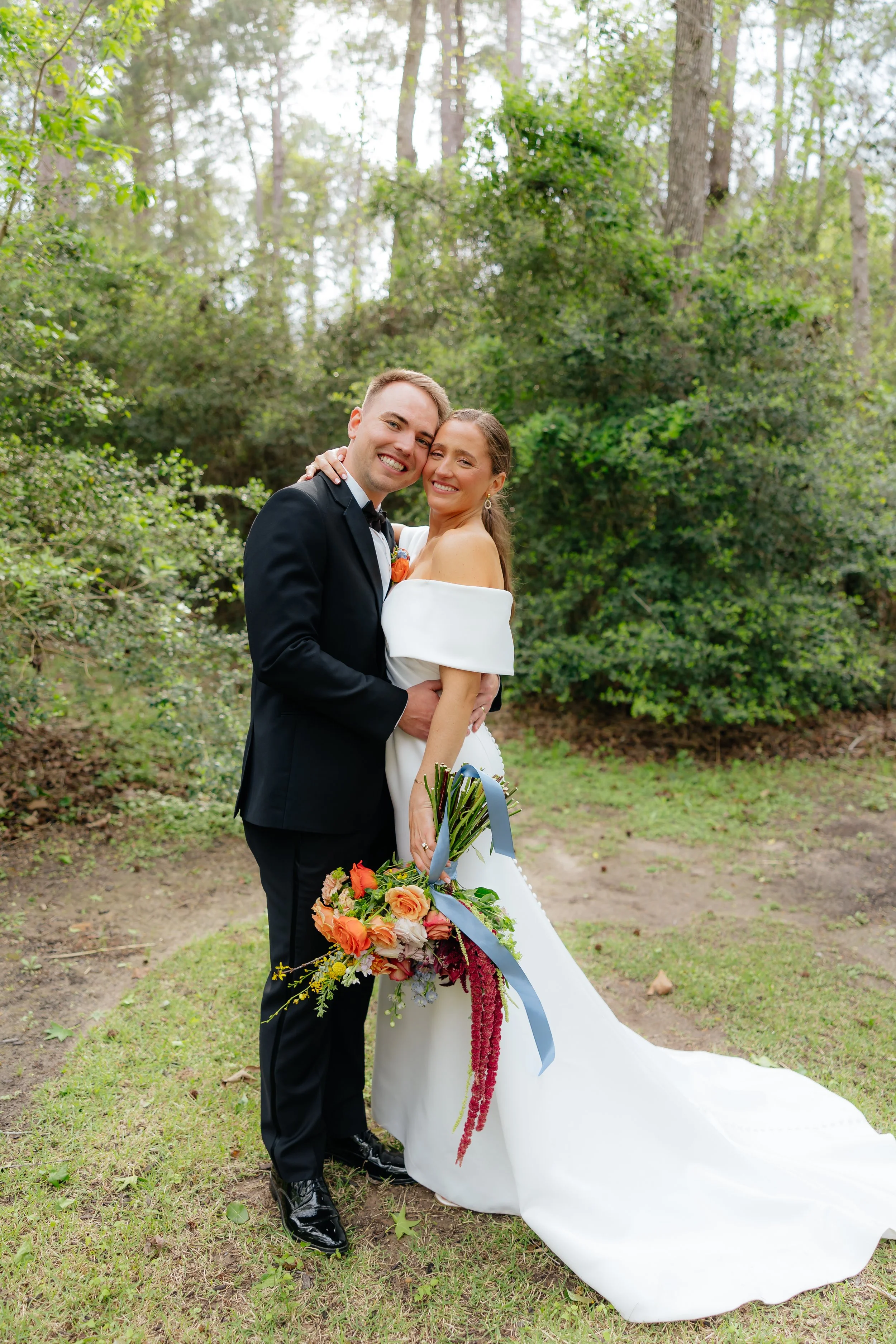 A happy bride and groom embracing outdoors in a wooded area, smiling at the camera, with the bride holding a colorful bouquet of flowers.