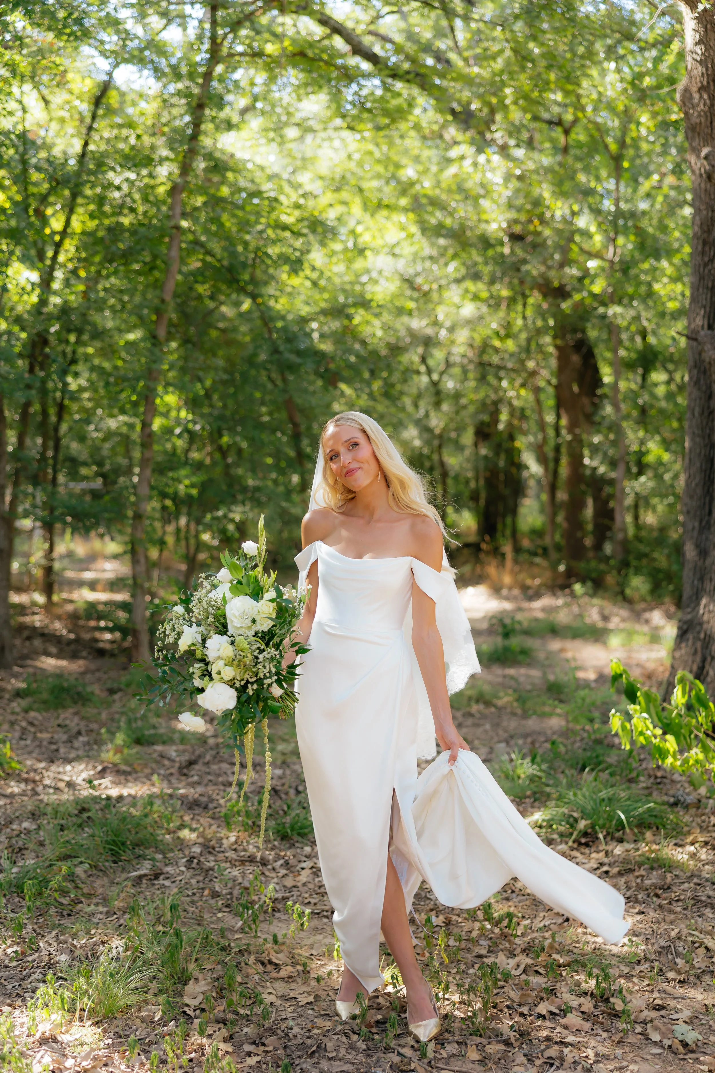 A bride with long blonde hair wearing a strapless white wedding gown, holding a large bouquet of white and green flowers, walking through a wooded area.