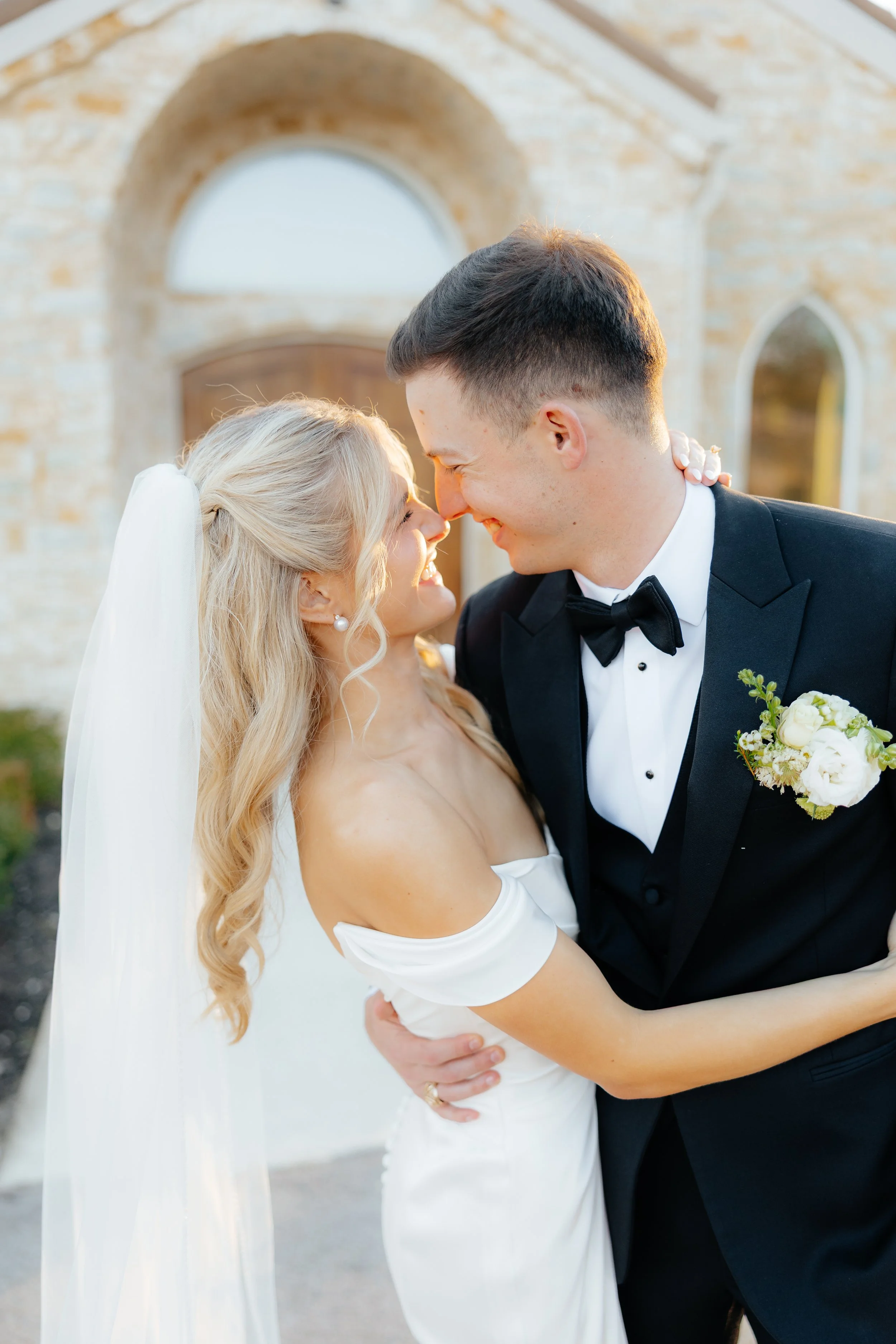 A newlywed couple smiling and touching foreheads outdoors at sunset, with a stone building in the background.