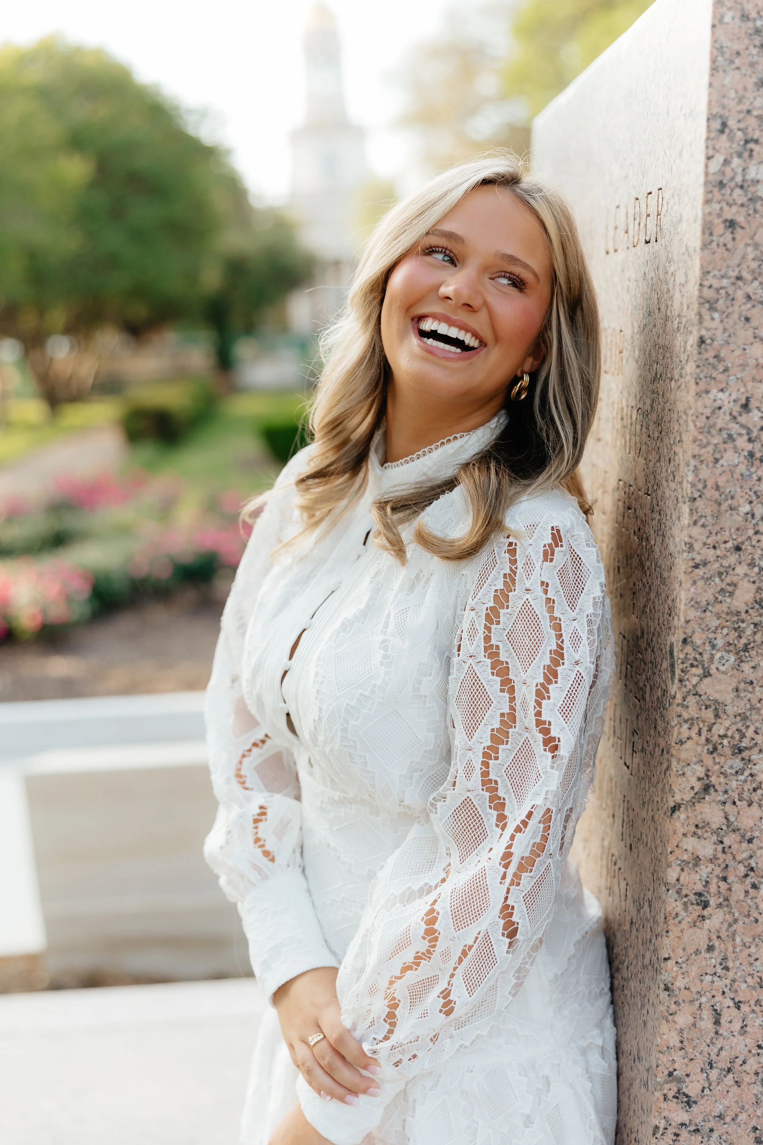 A woman with blonde hair wearing a white lace dress, smiling and leaning against a granite wall outdoors.