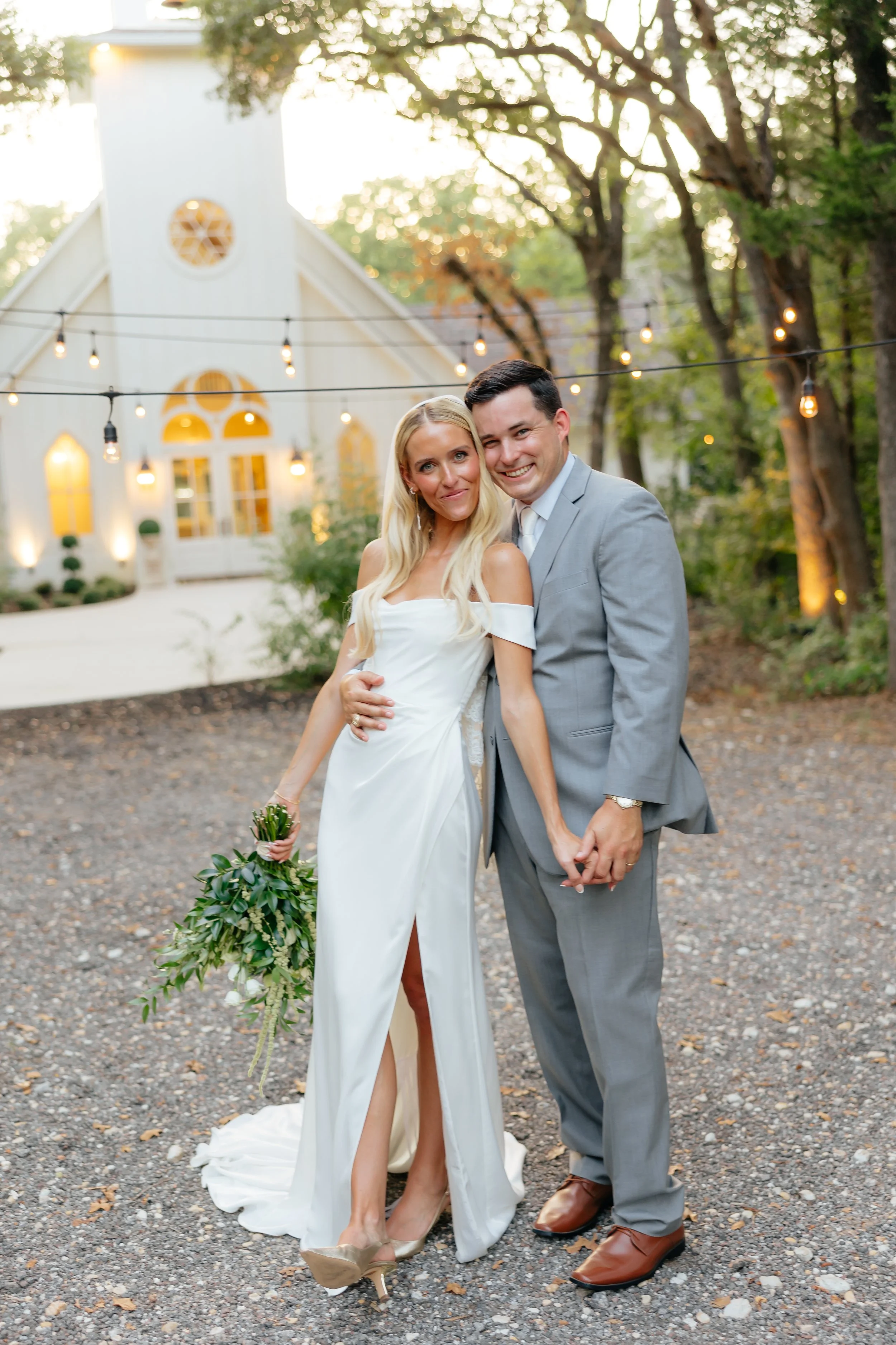 A newlywed couple smiling and holding hands outdoors during sunset, with a church building in the background and string lights above them.
