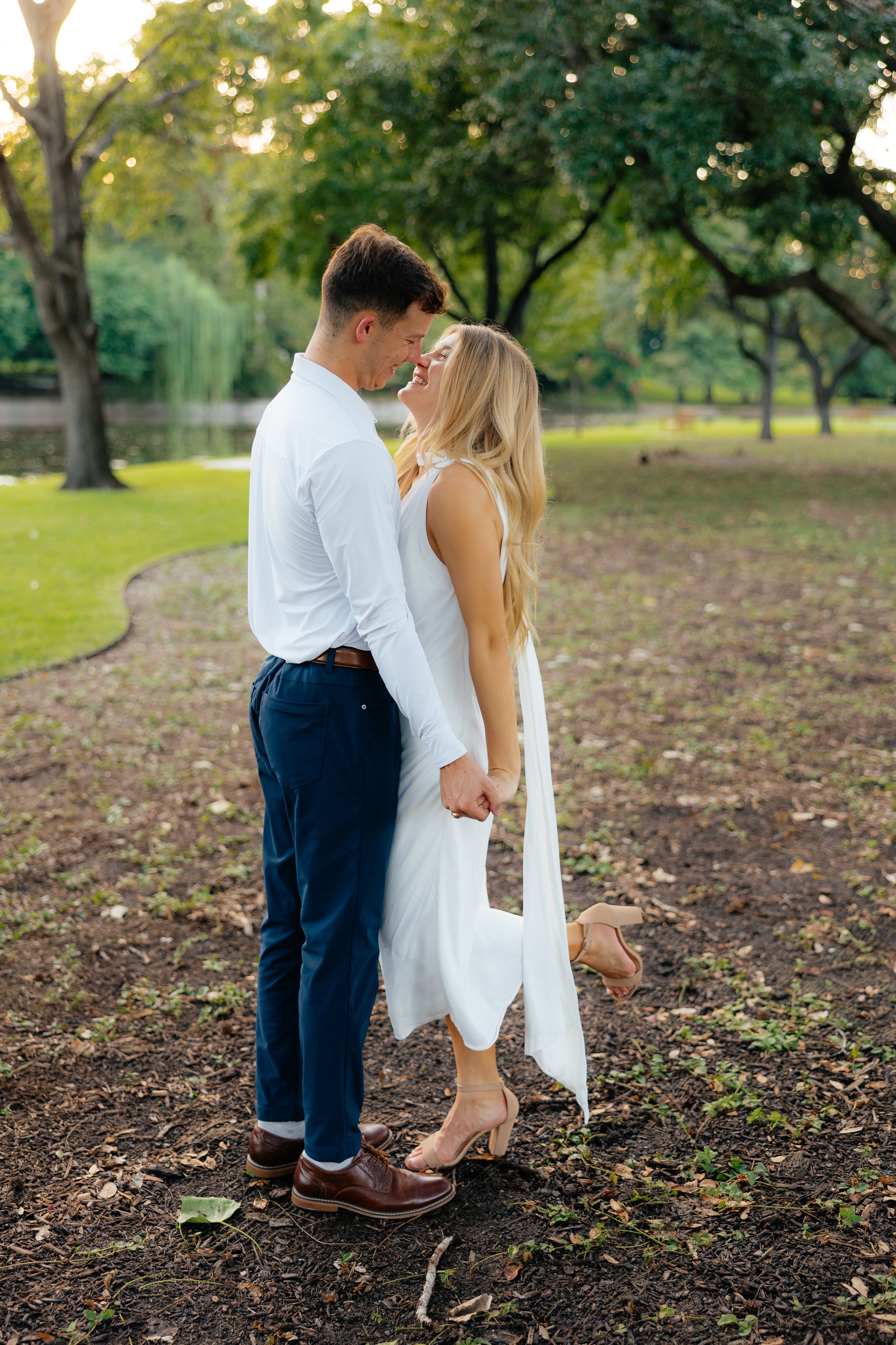 A young couple in formal attire, holding hands and smiling in a park with green trees and a pond in the background, during daylight.