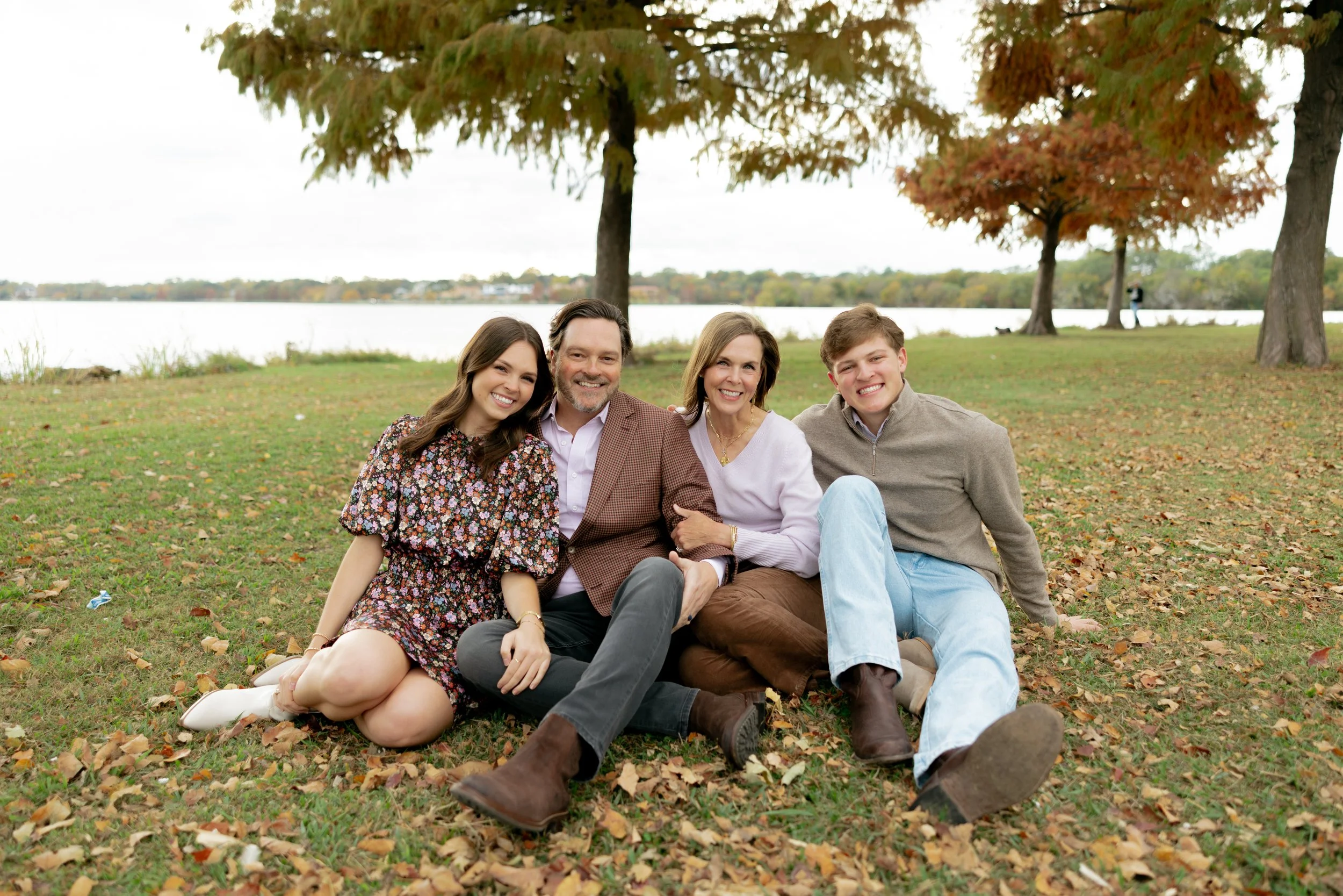 A happy family of four sitting on the grass in a park with autumn trees and a lake in the background.