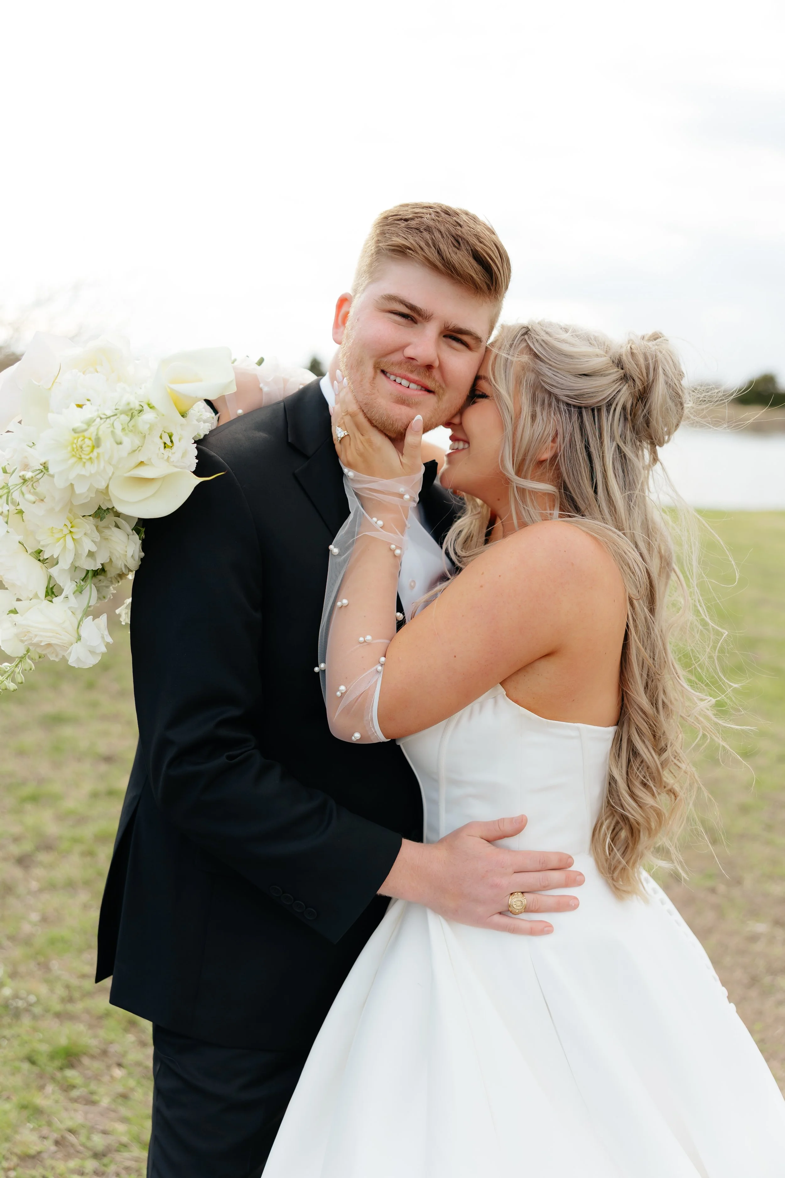 A newlywed couple is embracing outdoors by a body of water, with the bride holding a large bouquet and smiling as she leans into the groom. The groom is wearing a black suit and is smiling at the camera.