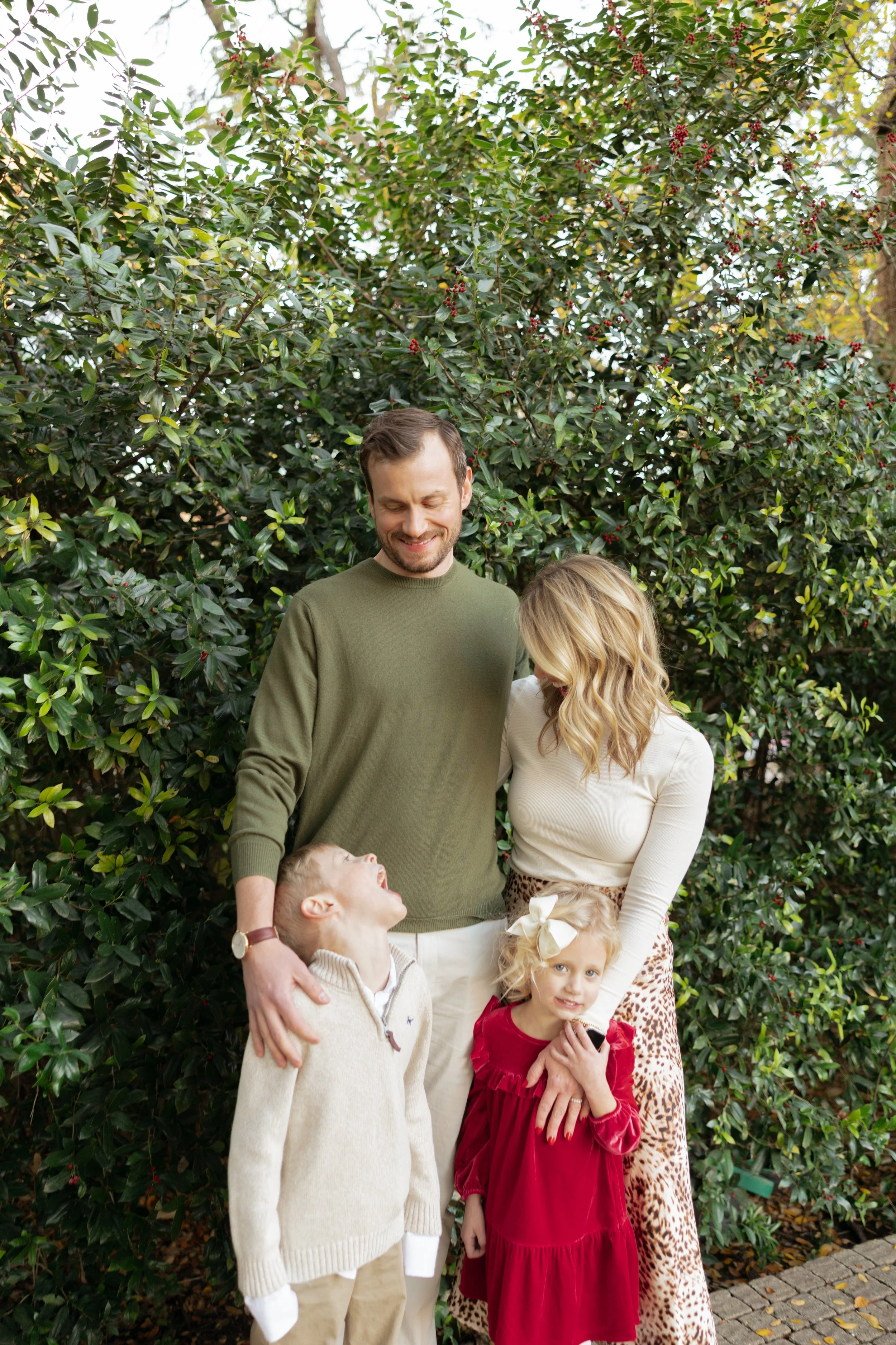 A family of four standing outdoors in front of a lush green shrub. The father, wearing a green sweater, smiles with eyes closed. The mother, in a cream top and leopard print skirt, looks down at their daughter, who is dressed in a red dress with a wh