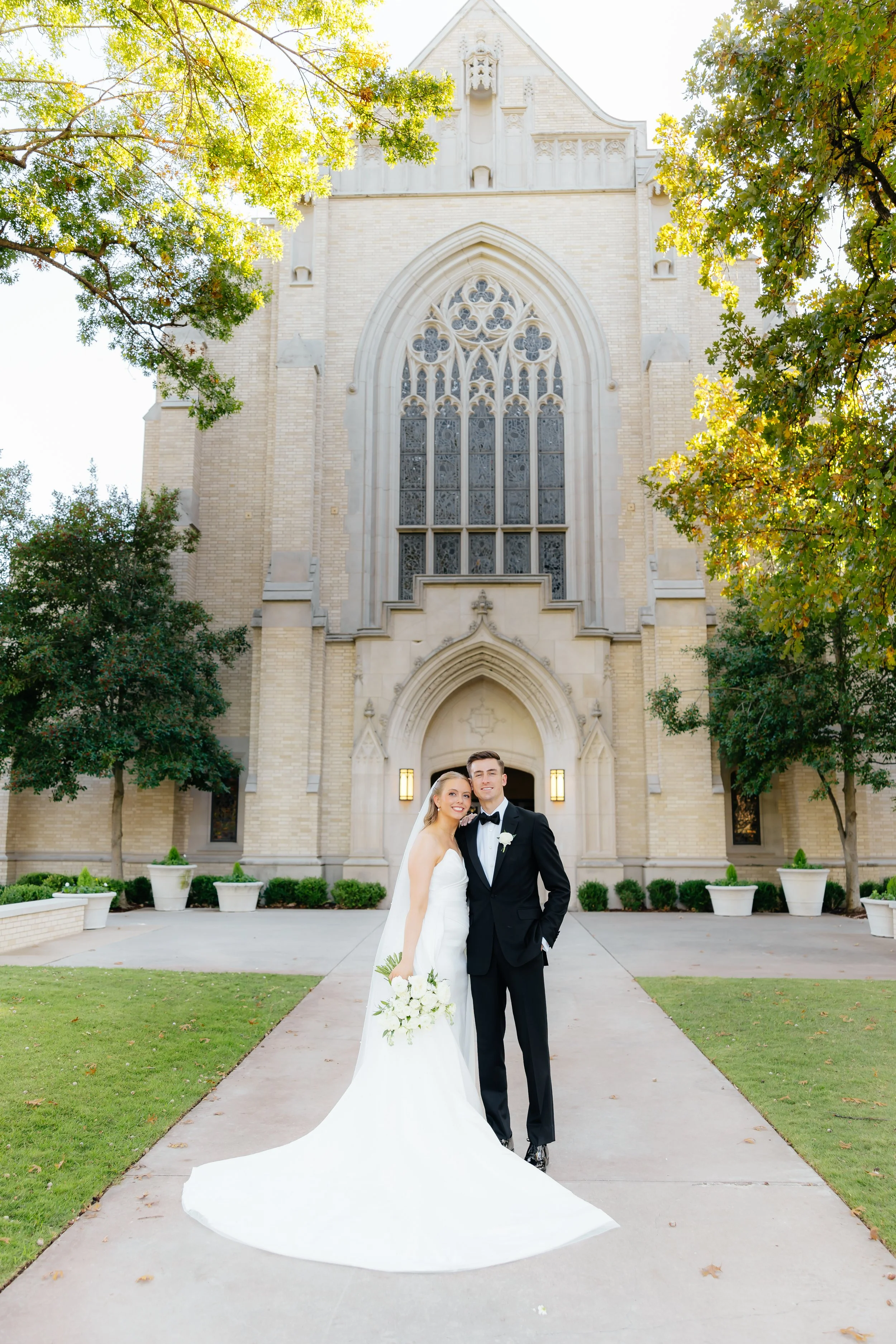A bride and groom in wedding attire standing in front of a large church building with stained glass windows, trees, and a pathway.