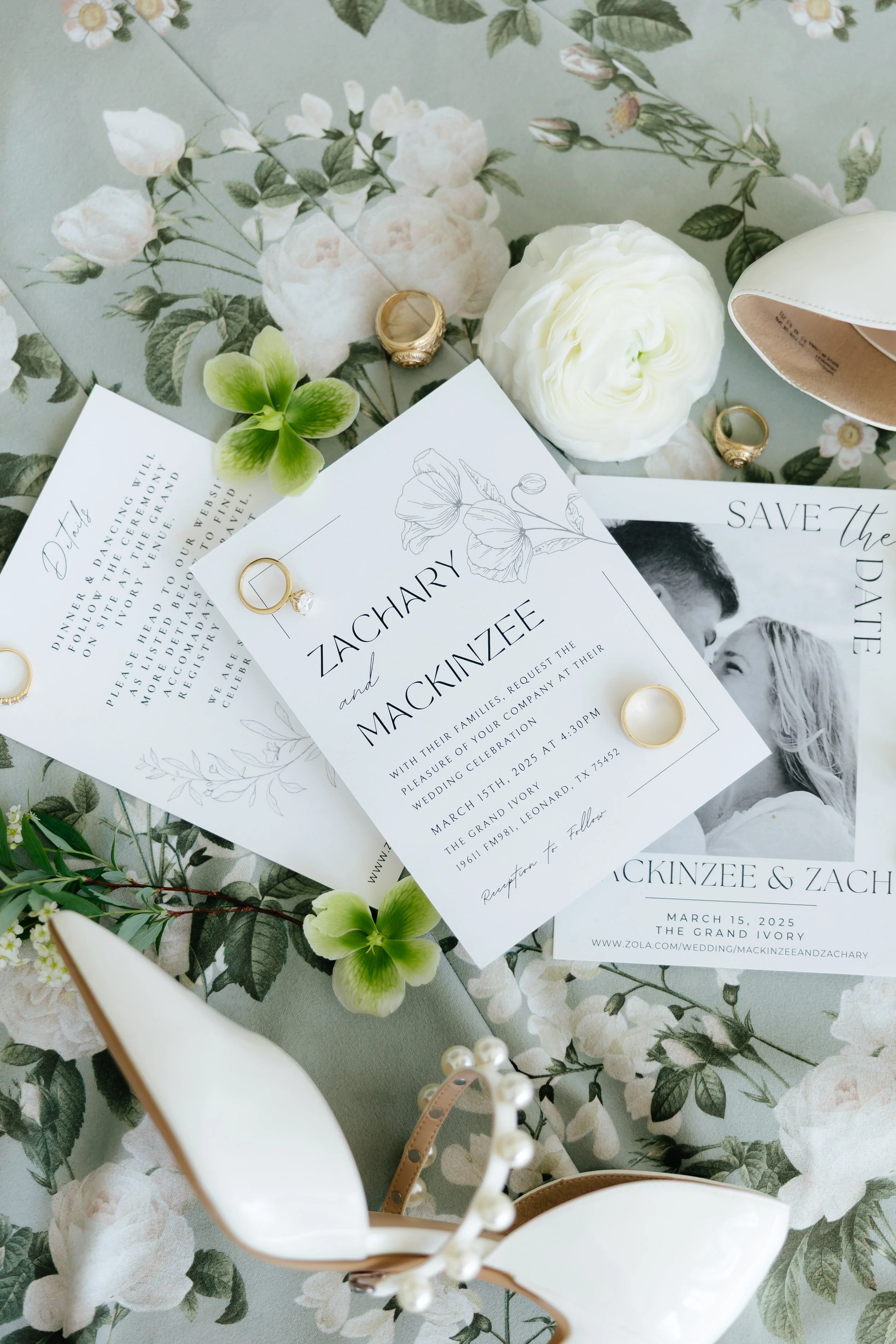 Wedding invitation suite on a floral tablecloth with floral decor, rings, white flower, and a pair of white high-heeled shoes.