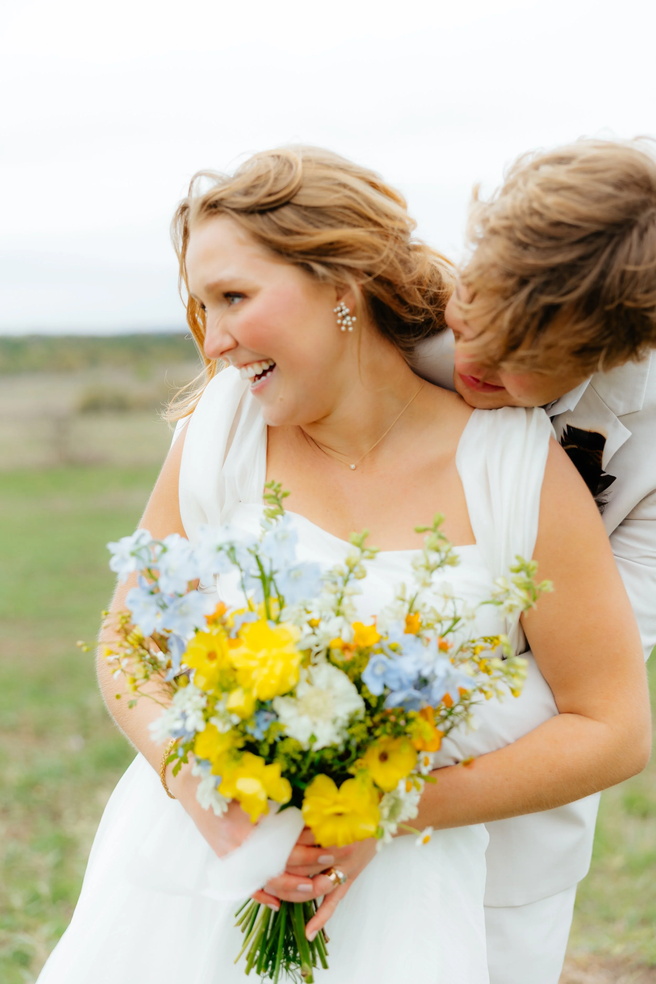 A joyful bride with wavy red hair smiling and holding a colorful bouquet of yellow and blue flowers, with a groom behind her in a white suit, outdoors in a grassy field.