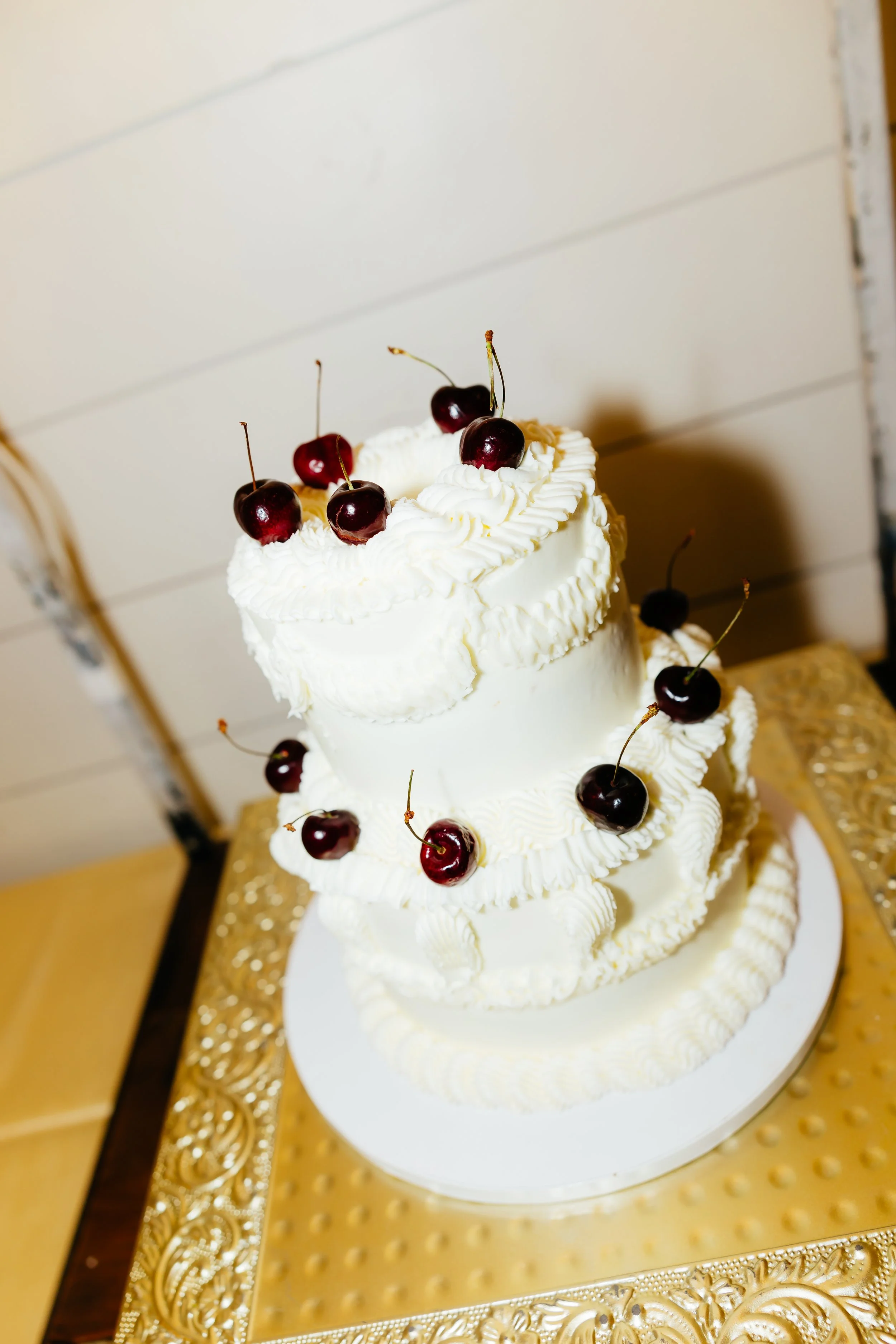 Three-tiered white wedding cake decorated with cherries and piped white frosting, placed on a gold-colored decorative table.