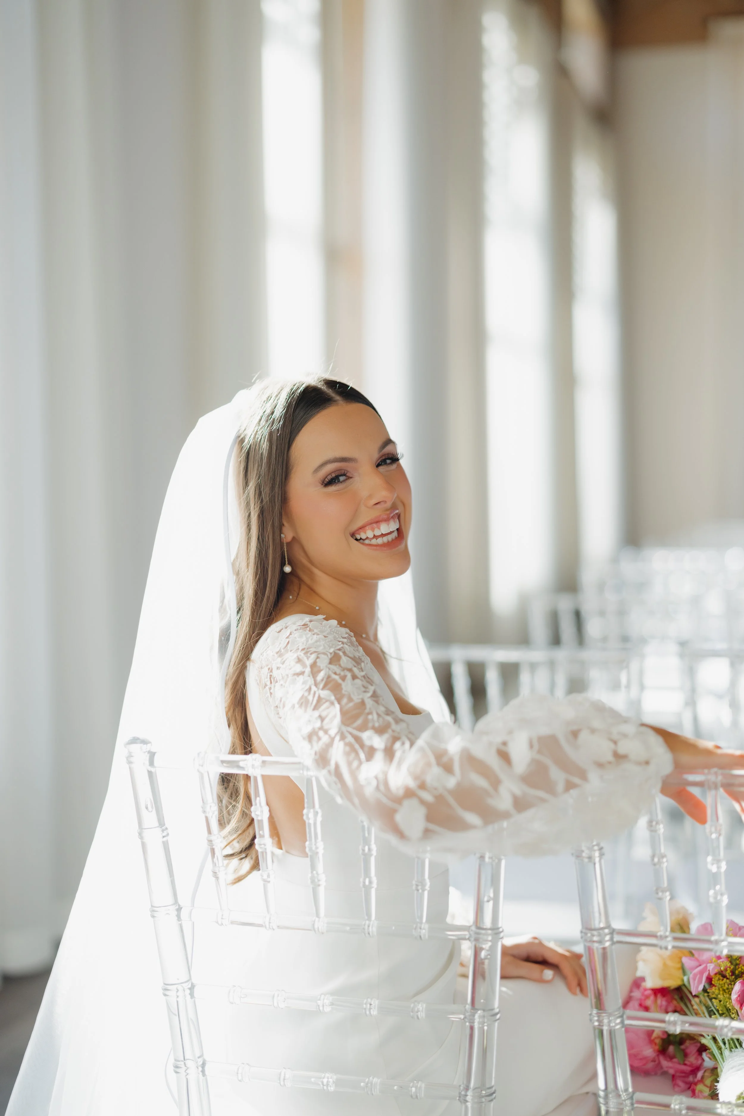 A smiling bride in a white wedding dress with lace sleeves, sitting on a transparent chair, with sunlight coming through windows in the background.