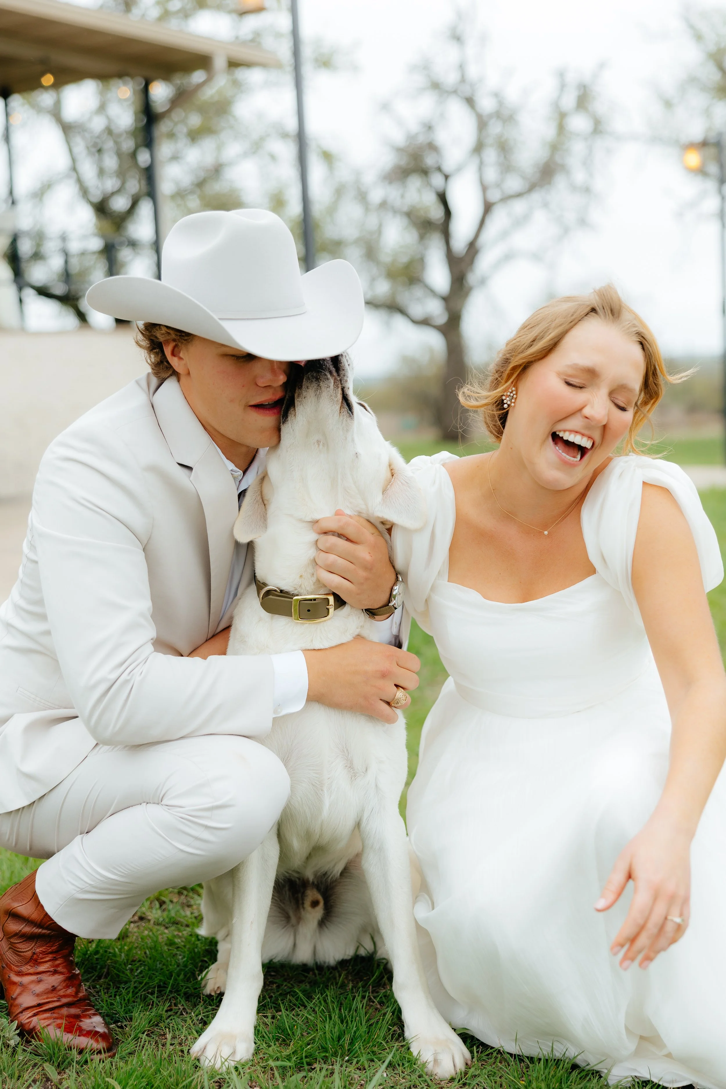 A young man in a white cowboy hat and suit and a woman in a white dress are sharing a moment with a happy baby goat outdoors on grass, with trees and a metal structure in the background.