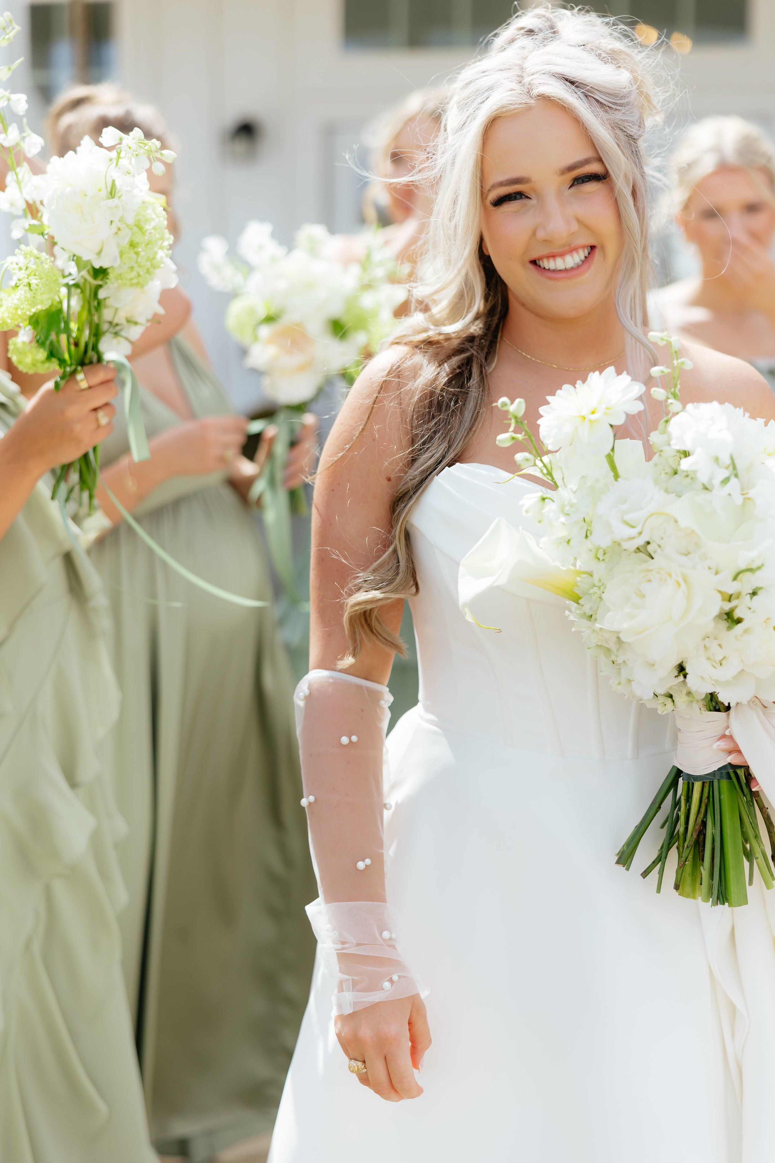A smiling bride in a white wedding gown holding a bouquet of white flowers, surrounded by bridesmaids in pastel dresses at an outdoor wedding ceremony.