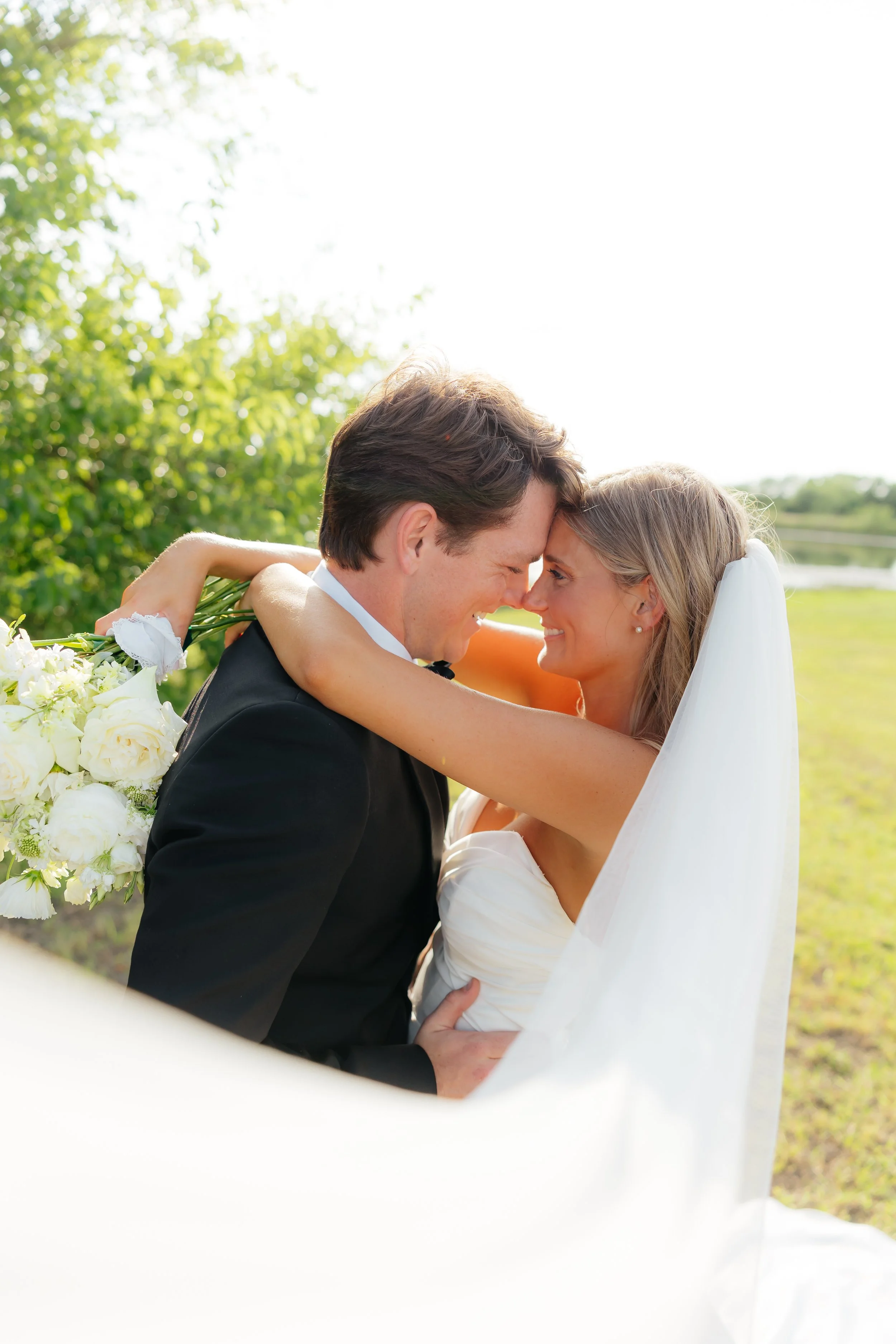 A bride and groom are embracing outdoors with their foreheads touching, smiling at each other. The bride is wearing a white wedding dress and veil, holding a bouquet of white flowers, and the groom is in a black tuxedo. The background features green 