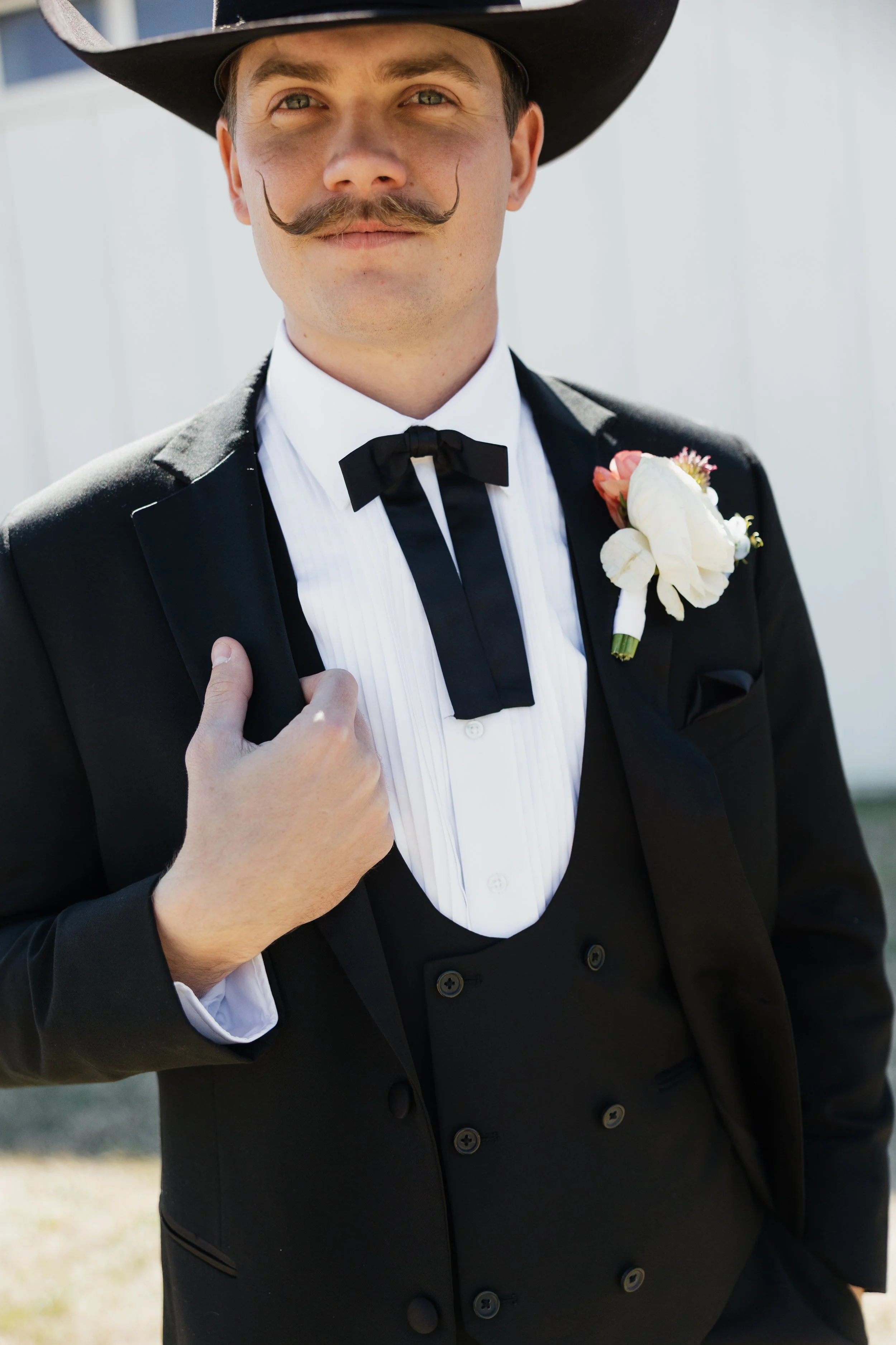A man dressed in a black tuxedo with a white shirt, black tie, and boutonniere, wearing a wide-brimmed hat, outdoors.