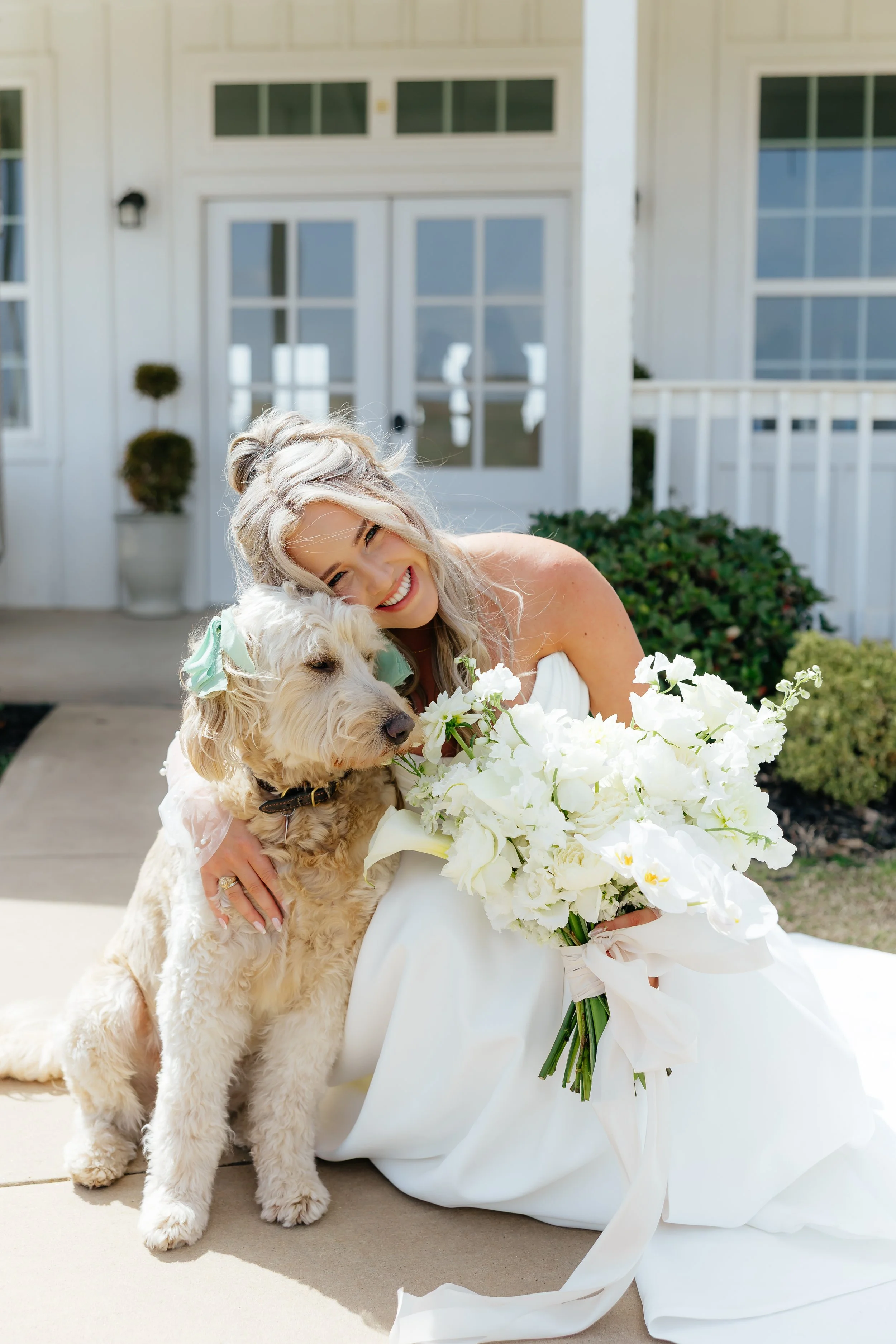 A smiling woman in a white dress hugging a golden doodle dog and holding a bouquet of white flowers, sitting outside in front of a white house with glass doors and windows.