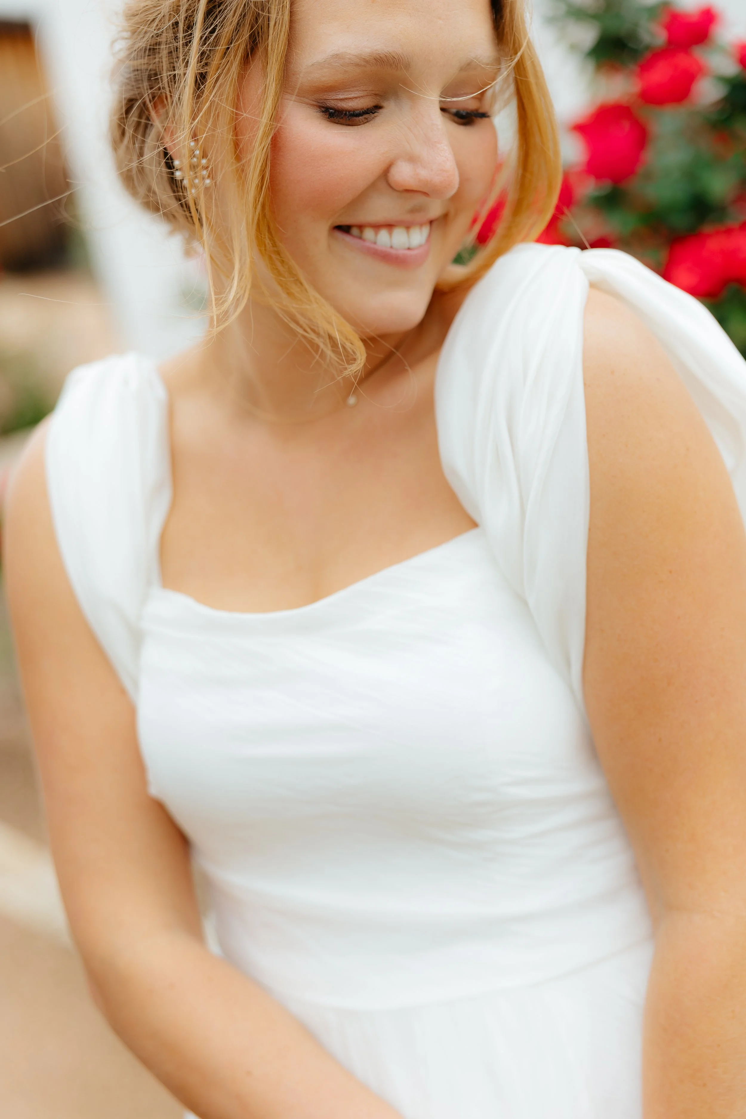 A smiling woman with blonde hair, wearing a white dress with puffed sleeves, is looking down and to her side. She is outdoors with red flowers in the blurred background.