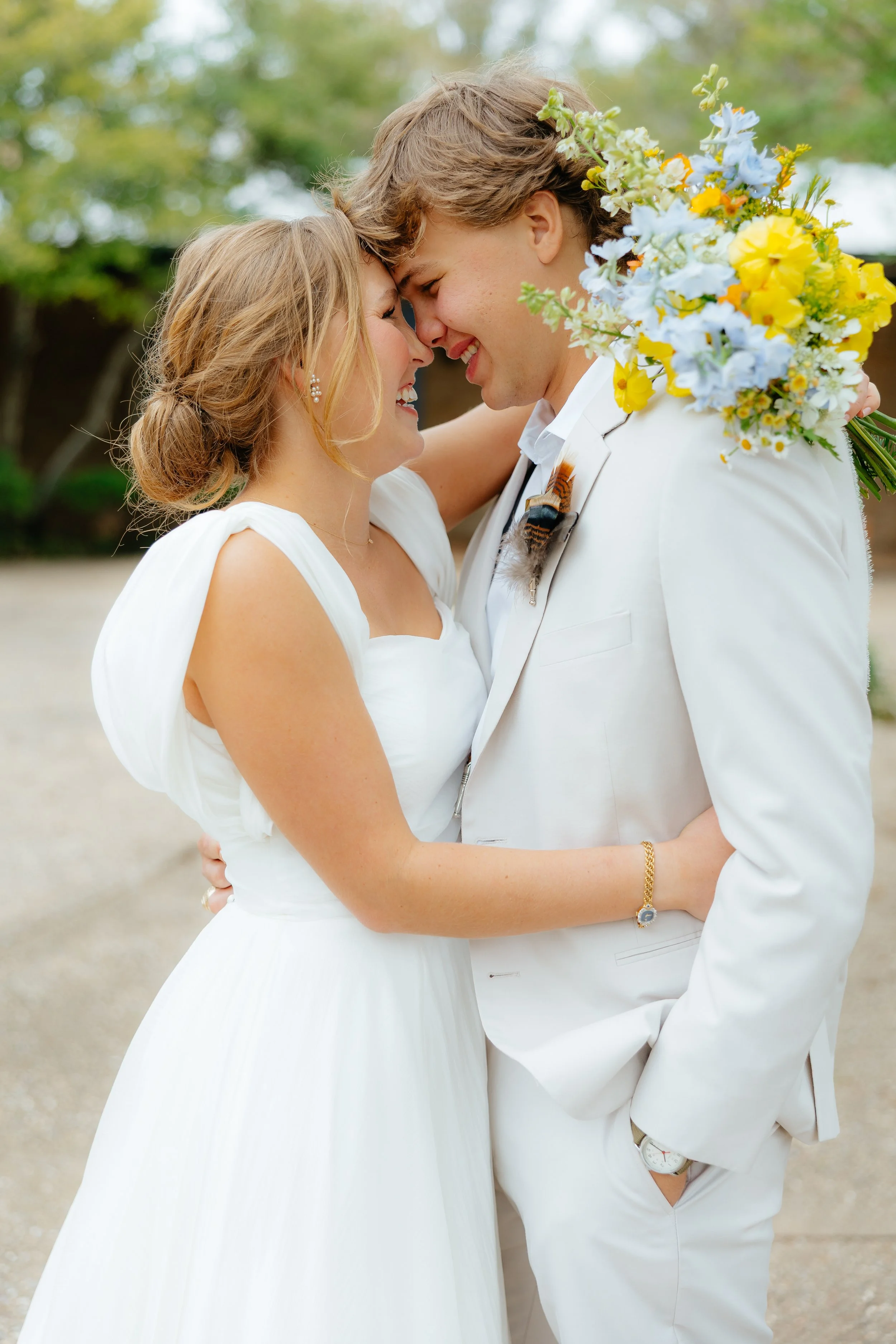 A bride and groom in wedding attire smiling and touching foreheads outdoors, with the groom holding a large colorful bouquet.